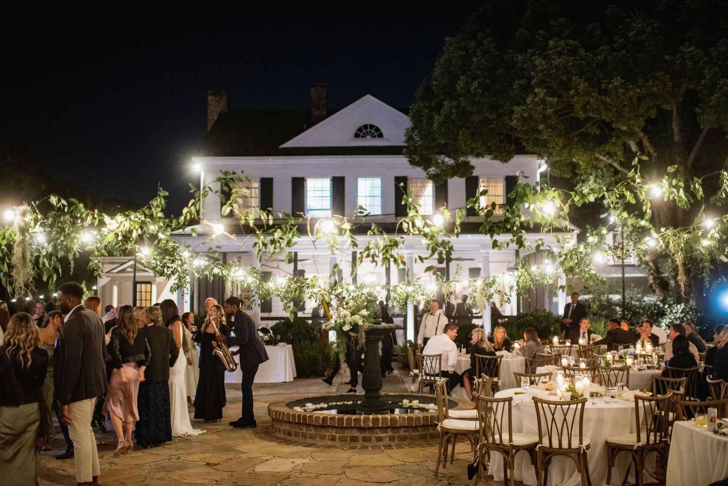 Outdoor evening wedding reception with string lights illuminating tables and guests under trees at a rustic venue.