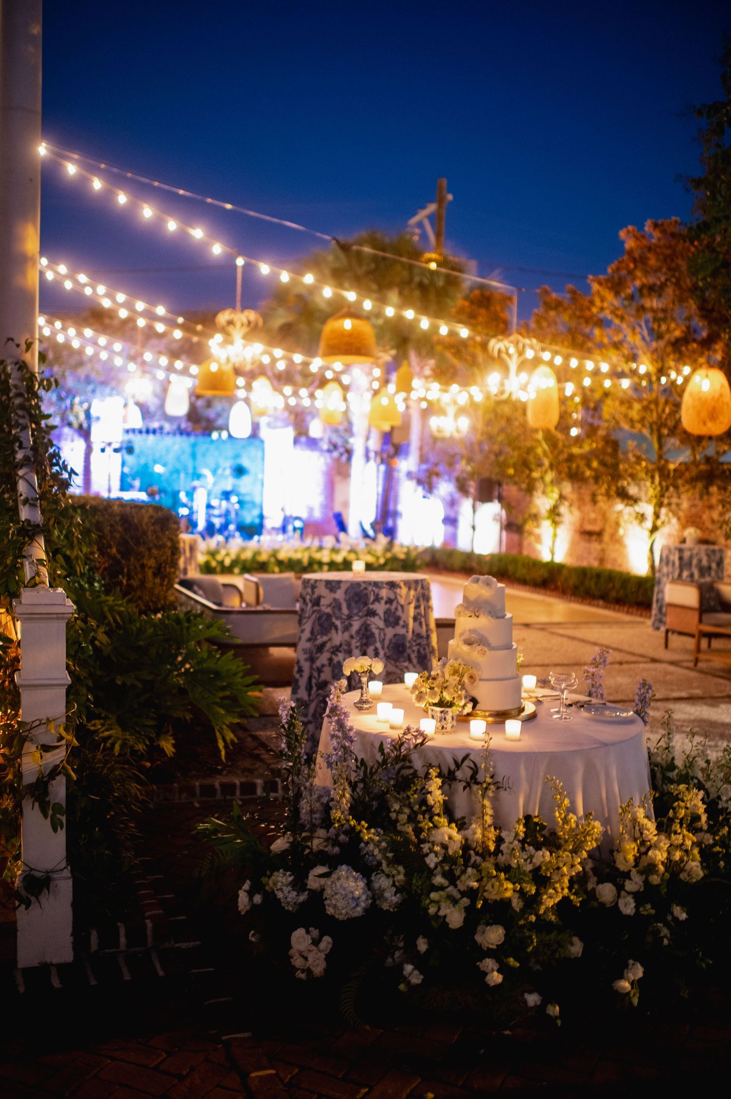 Elegant outdoor wedding reception setup at dusk with string lights and white floral arrangements on tables.