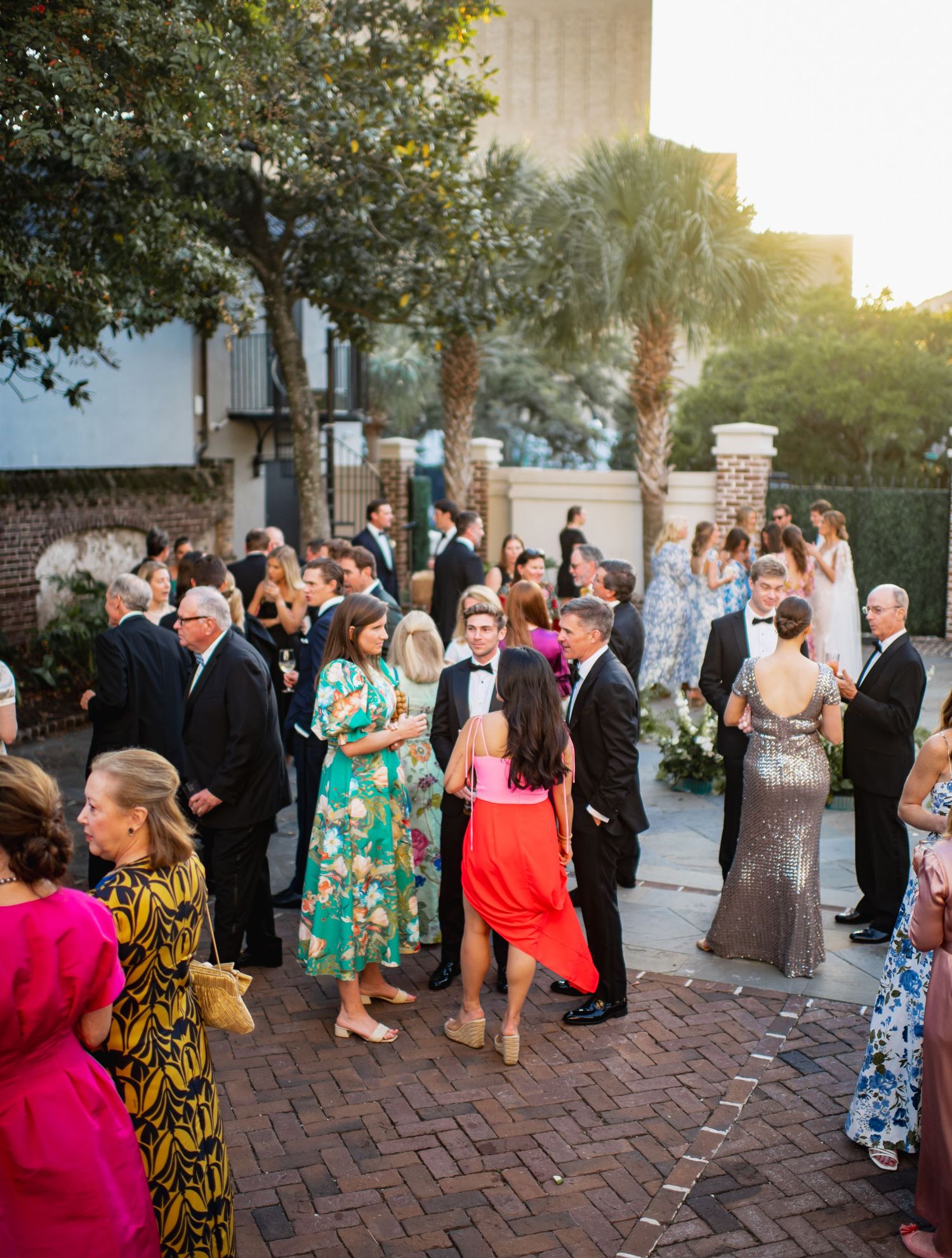Wedding guests gather at an outdoor cocktail reception on a brick patio with palm trees at sunset.