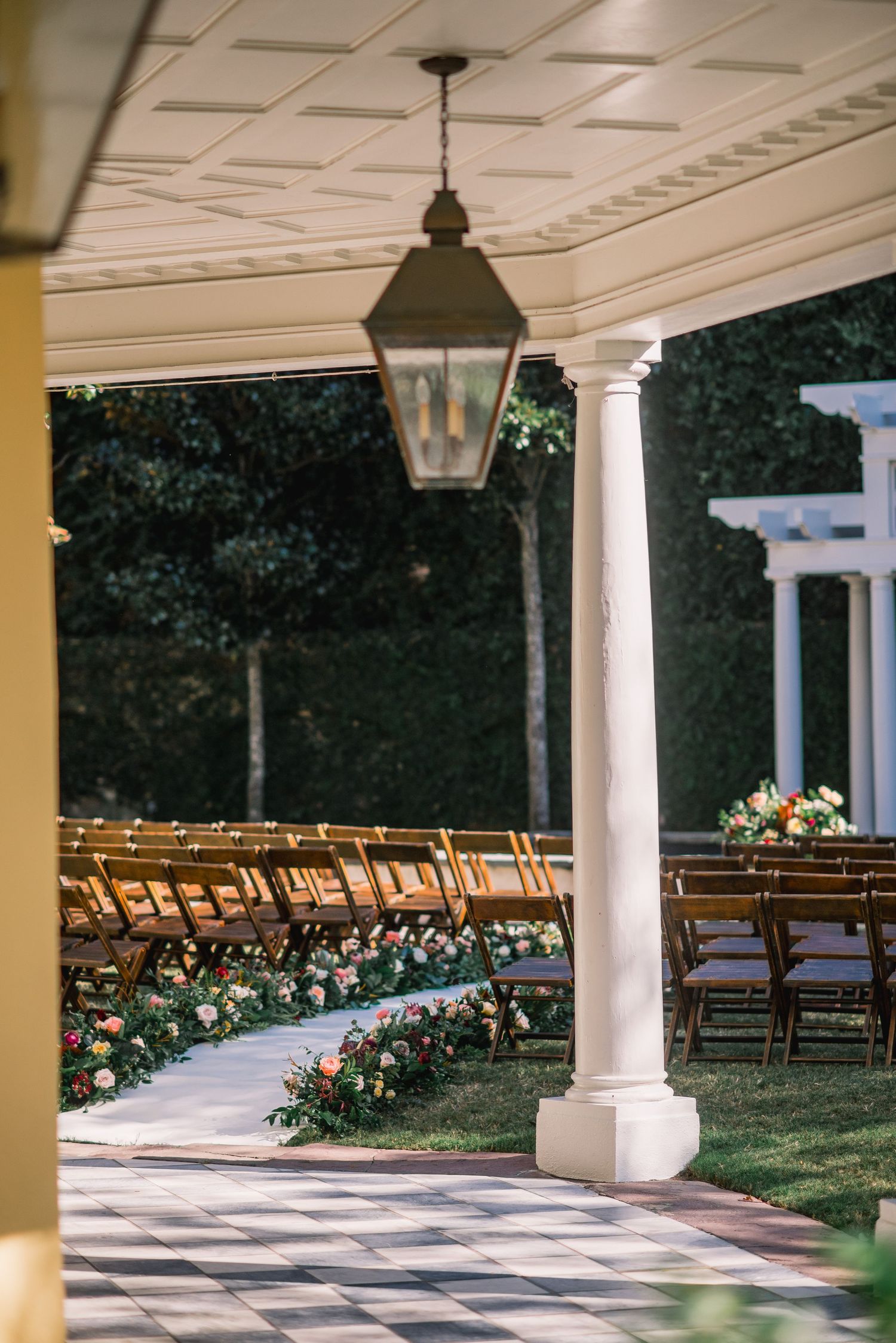 Curved outdoor wedding ceremony setup with wooden chairs and floral arrangements under a columned white portico.