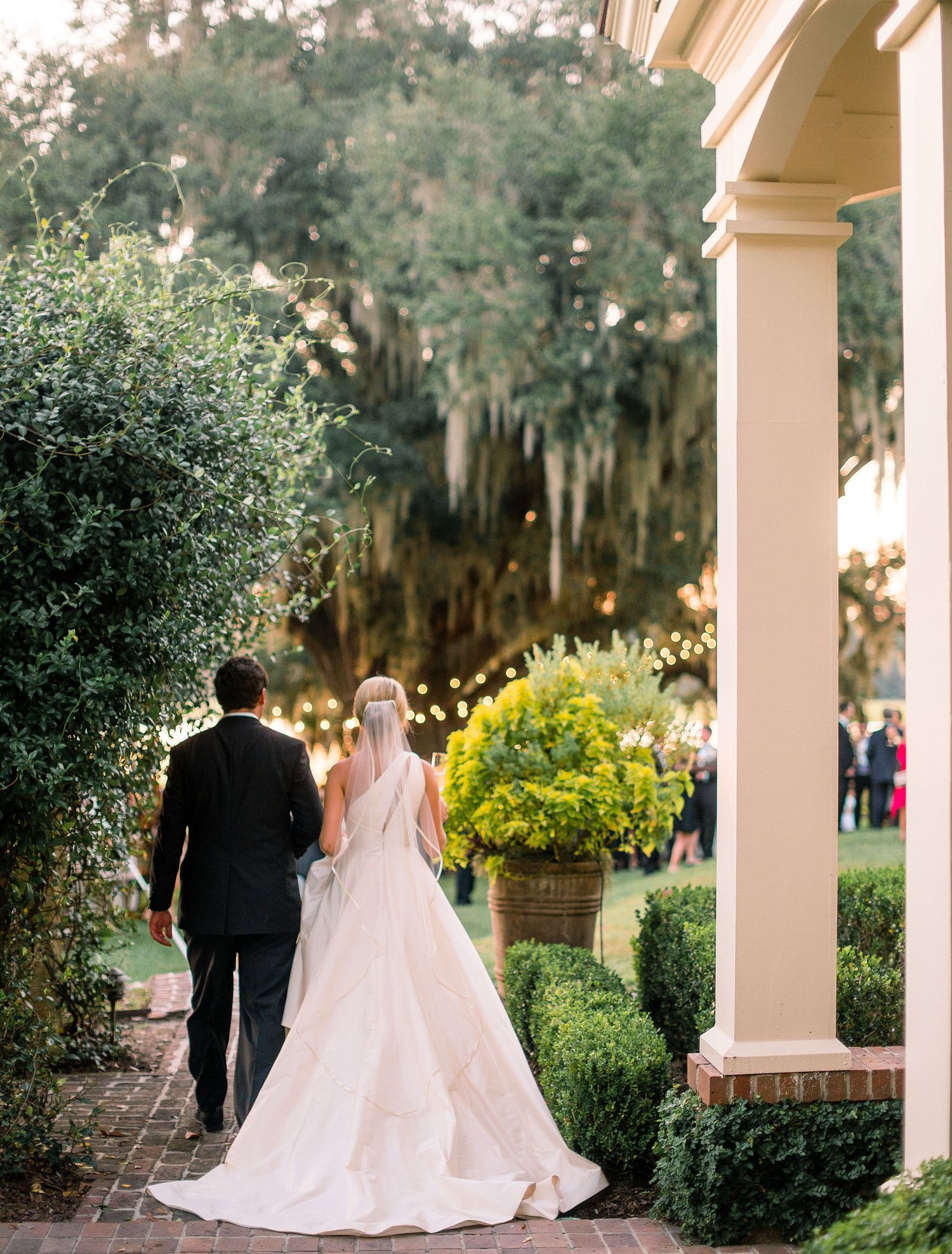 A couple in formal wedding attire walks along a covered walkway with Spanish moss-draped trees in the background.