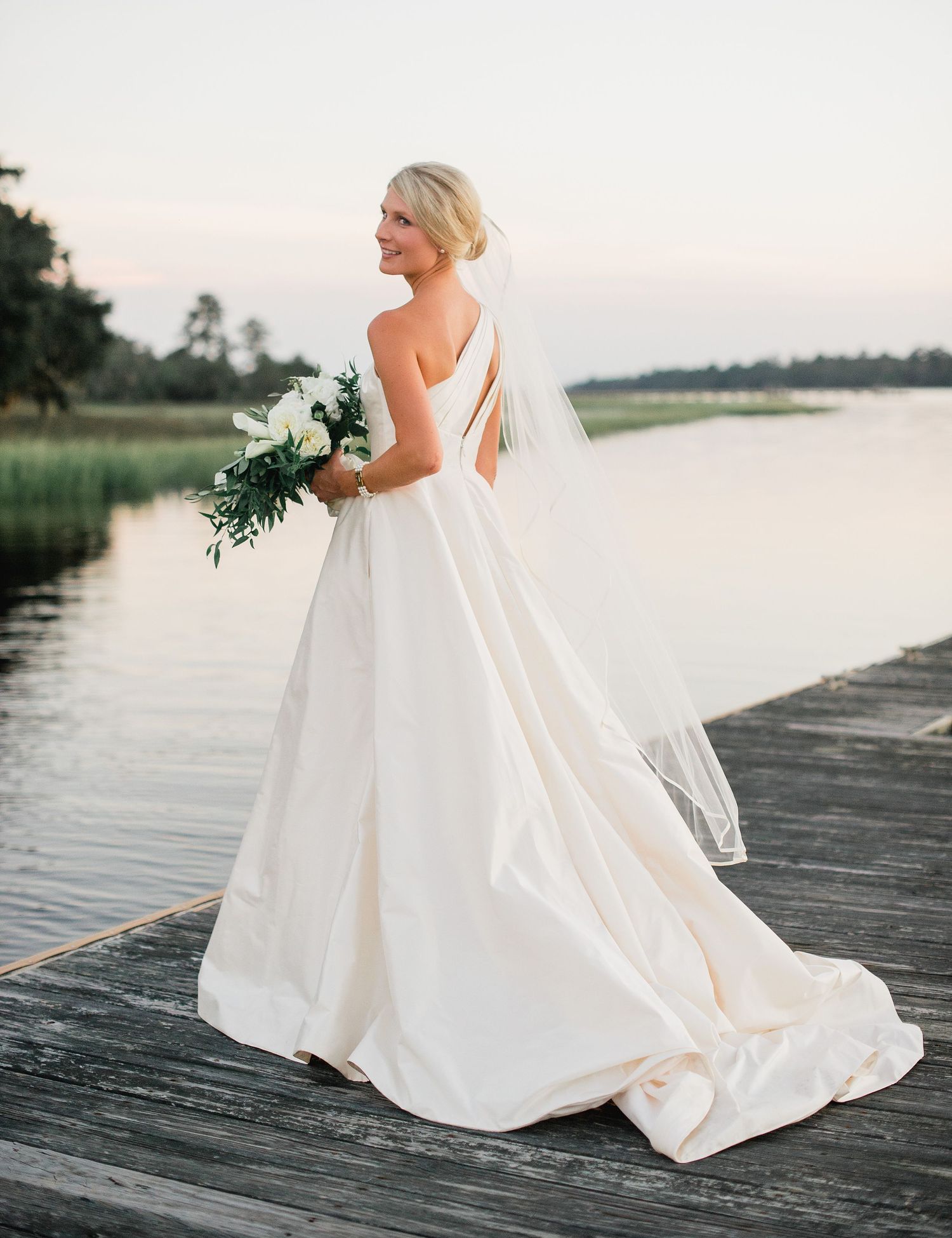 A bride in a flowing white gown stands gracefully on a wooden dock by a serene lake during sunset.