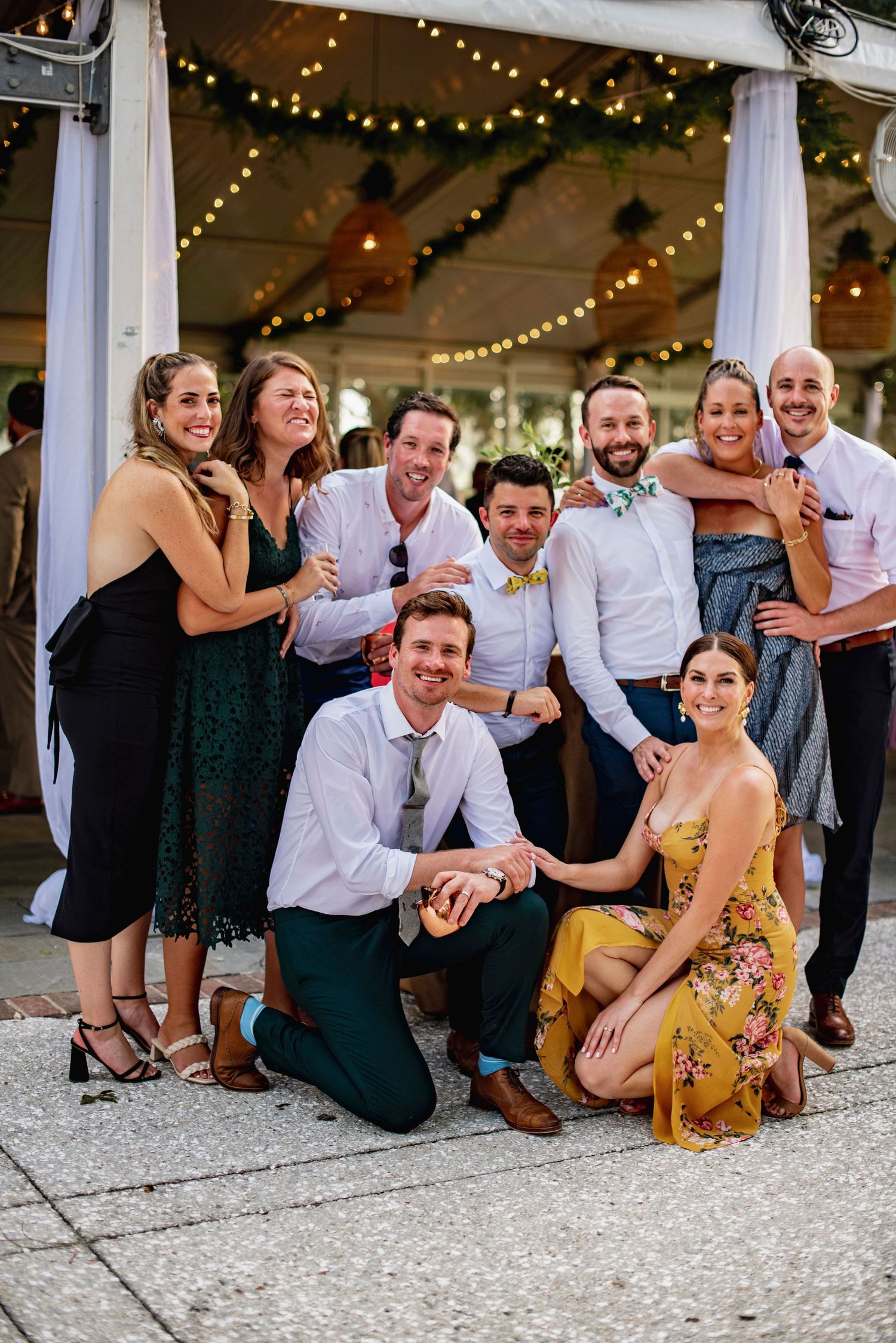 A group of wedding guests in formal attire pose together on the ground for a fun photo under string lights.