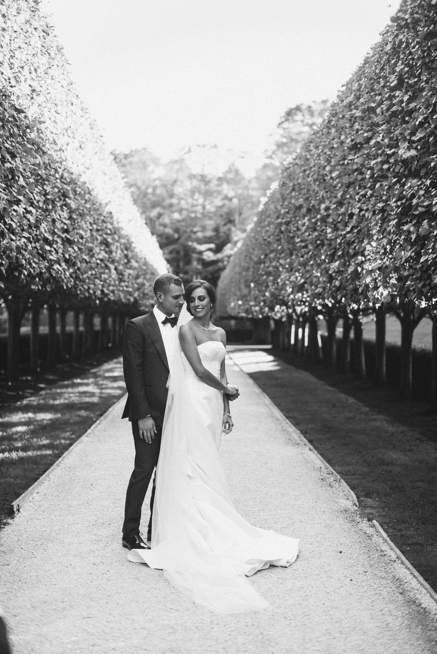 A black and white wedding photo shows a couple walking down a tree-lined pathway in an orchard setting.