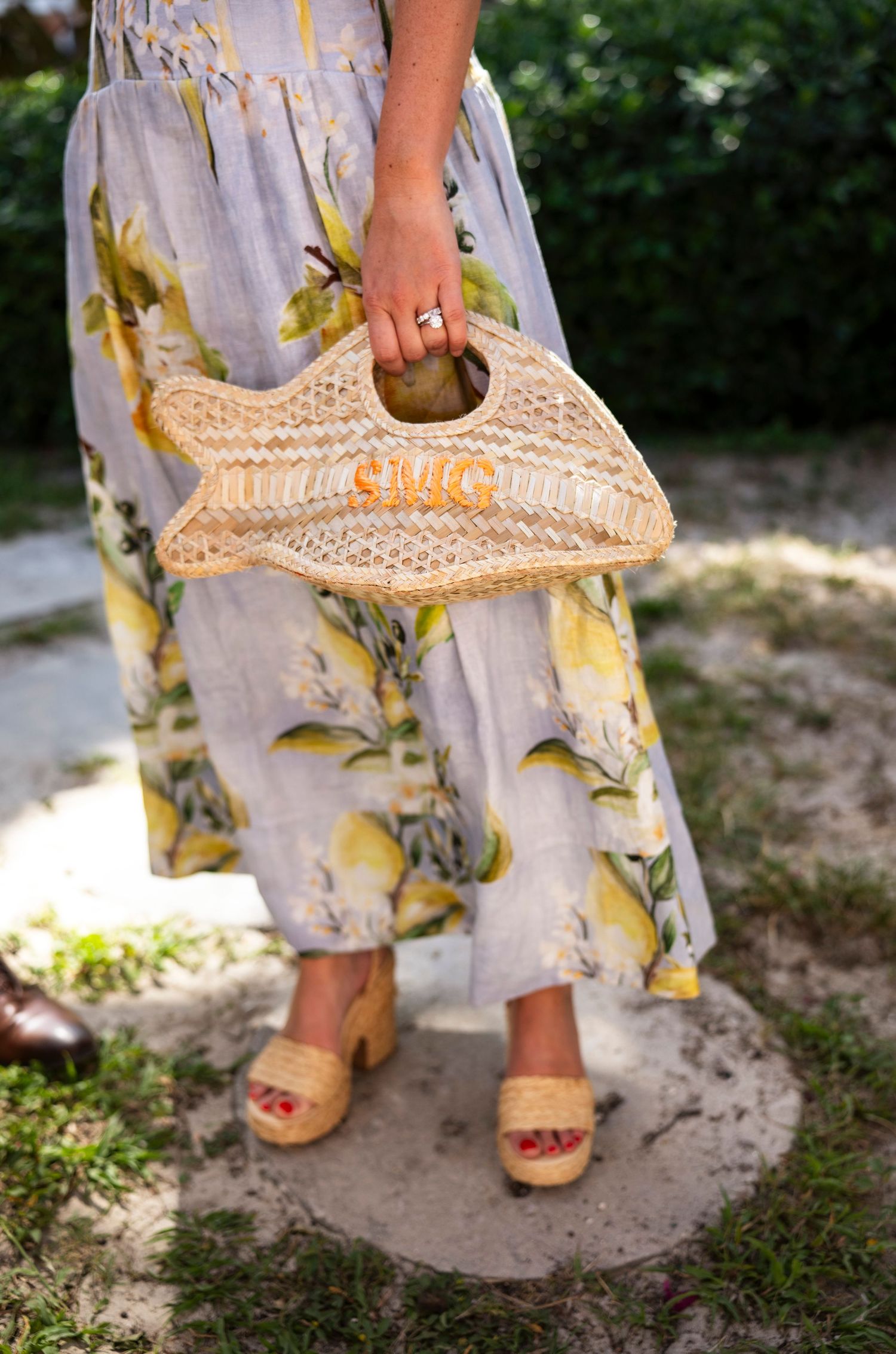 Person wearing floral dress holds straw beach bag while standing on garden path.