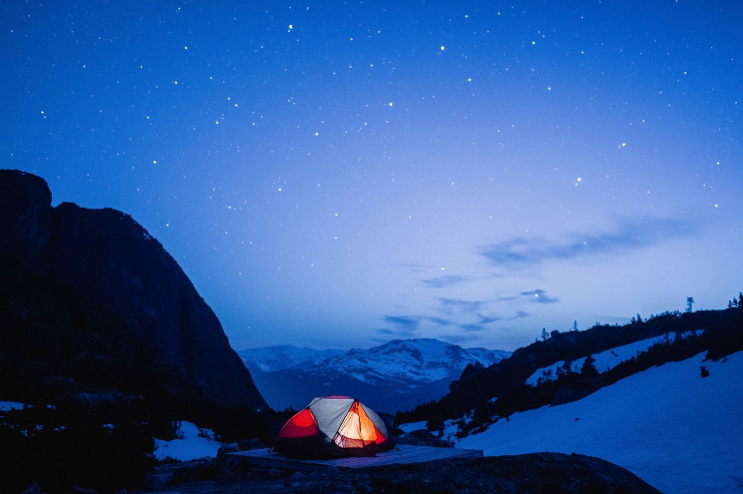 Illuminated camping tent glows against starry night sky in mountain landscape with silhouetted peaks.