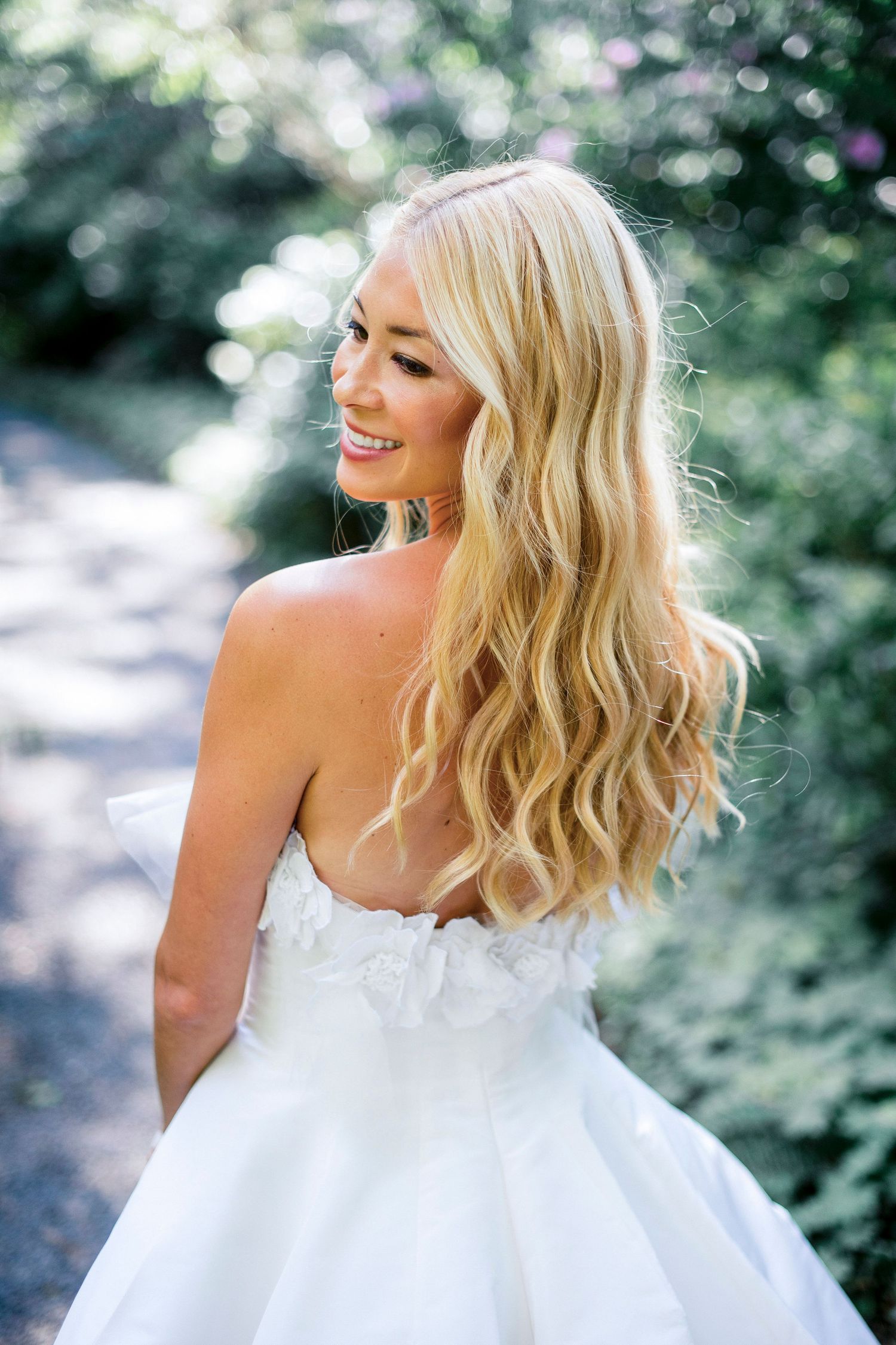 Bride in strapless white gown smiles while posing in garden setting.