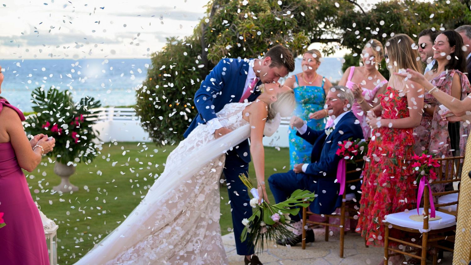 Wedding guests shower a couple with confetti during an outdoor ceremony by the water on a sunny day.