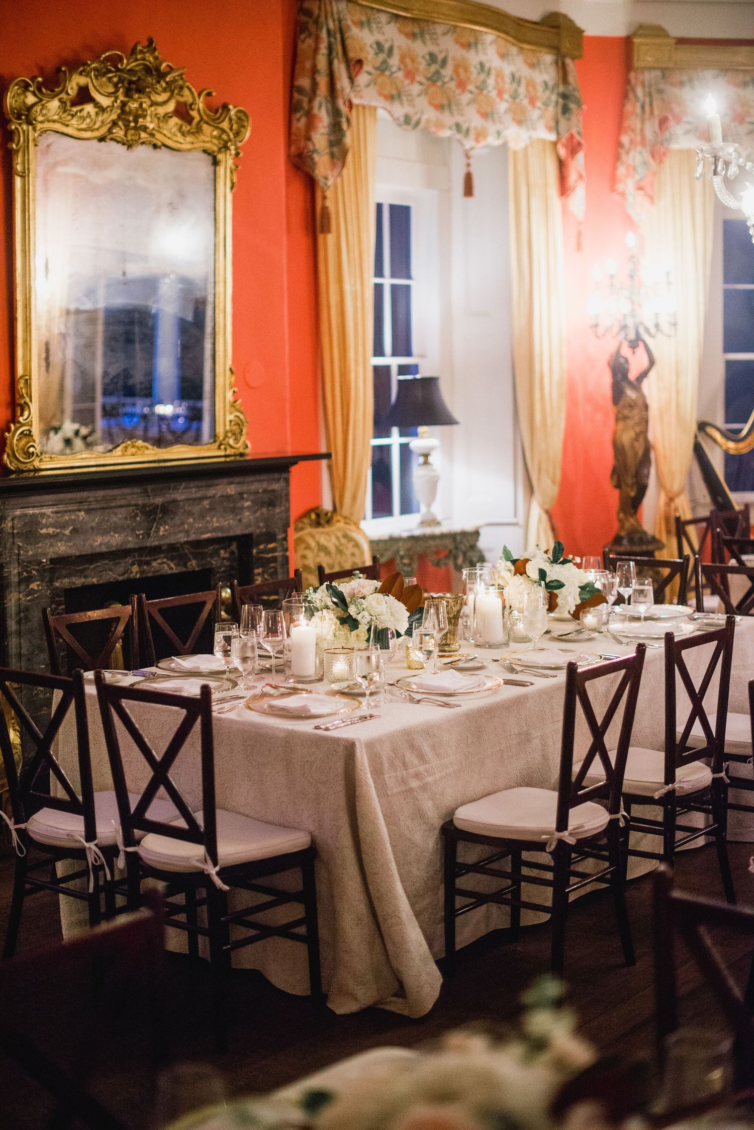 Elegant dining tables set with white linens and candles in a formal red-walled room with ornate gold-framed mirrors.