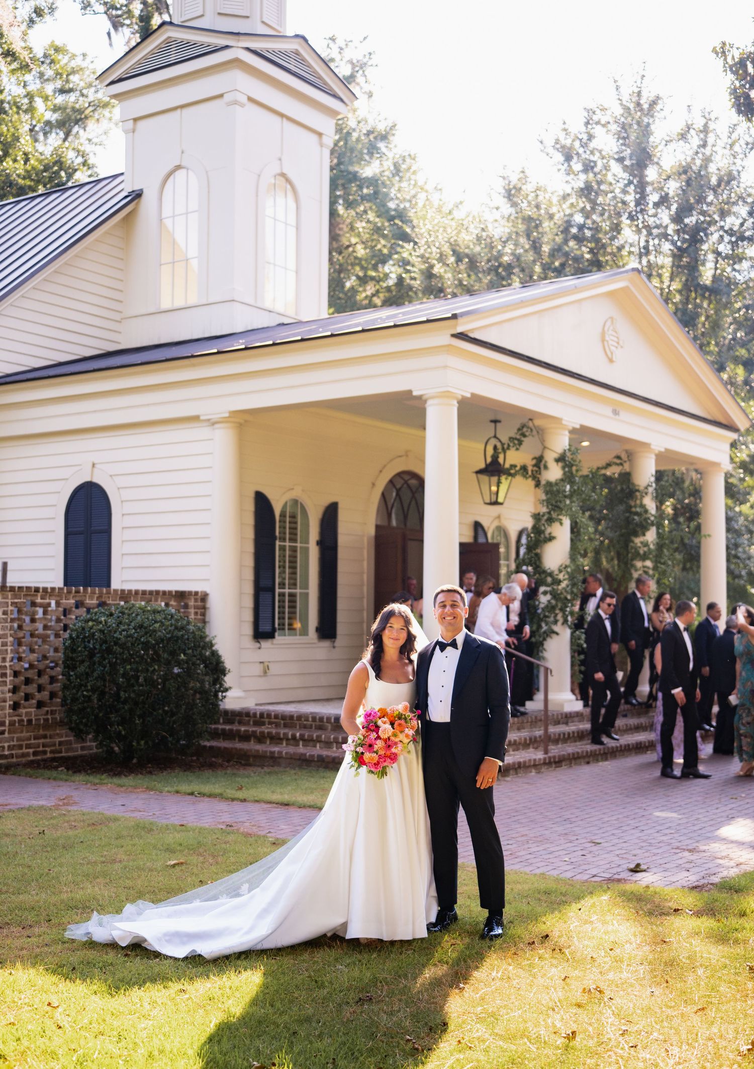 A wedding ceremony takes place at a charming white chapel with a covered porch and brick foundation on a sunny day.