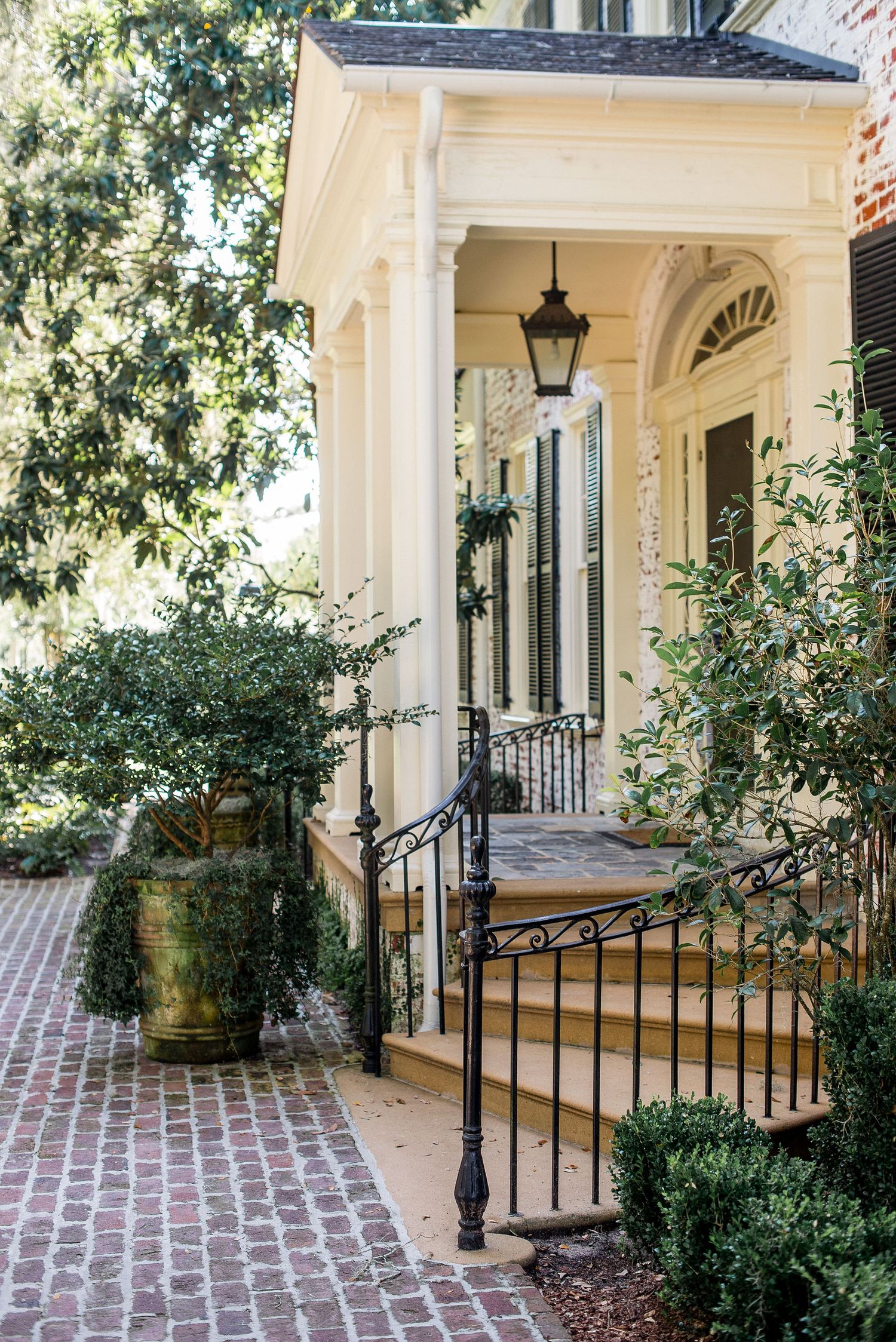 Brick walkway leads to a classic Southern porch with white columns, iron railings, and potted plants in natural light.