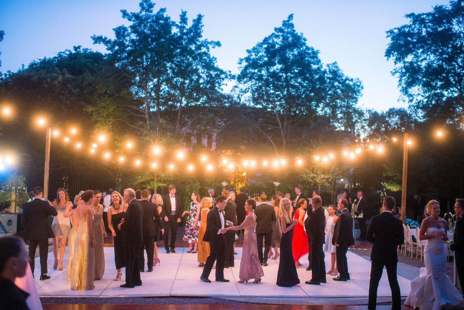 Wedding guests dance together under twinkling string lights at an outdoor evening reception.