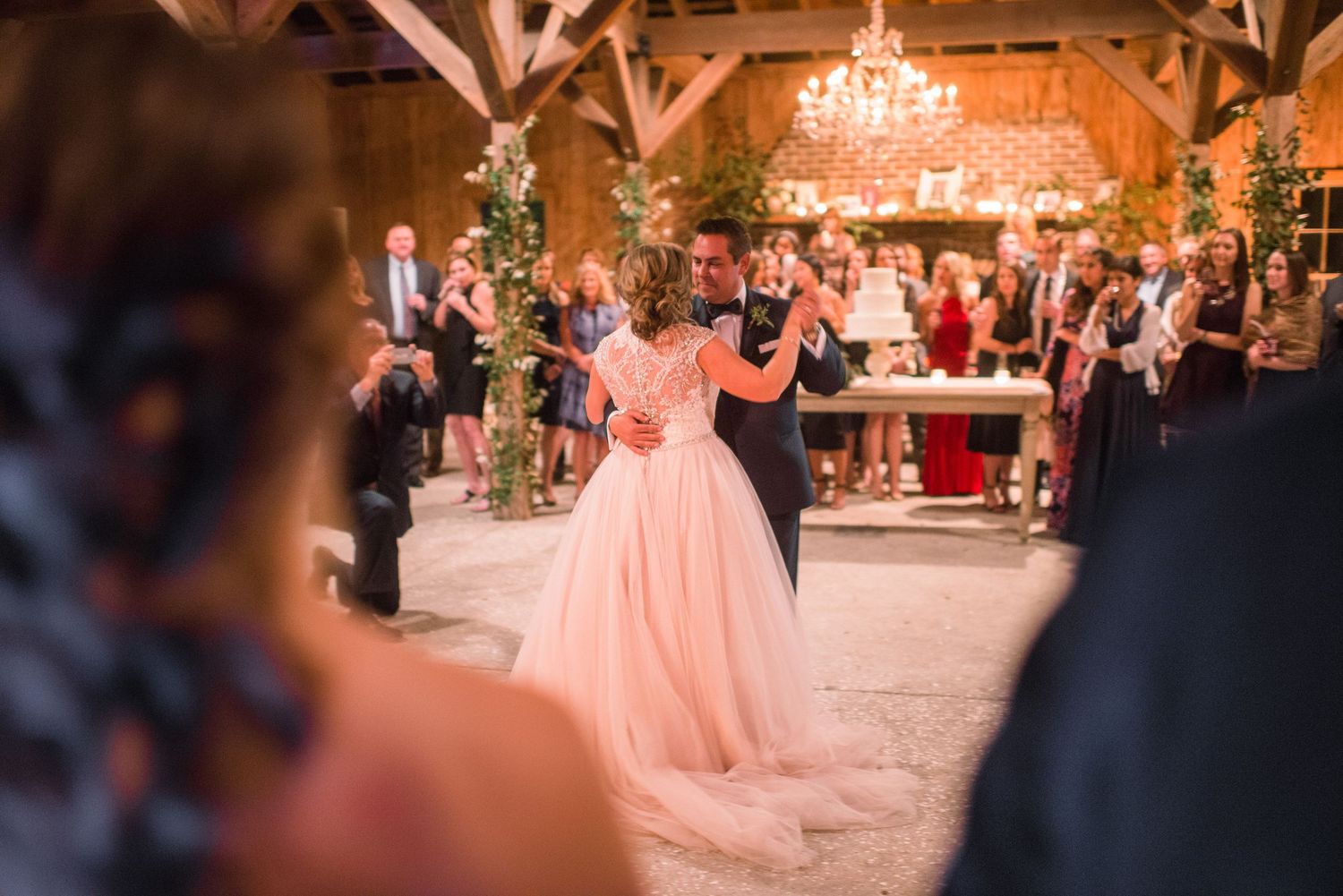 A couple shares their first dance at a rustic barn wedding reception with a pink tulle gown and crystal chandelier overhead.