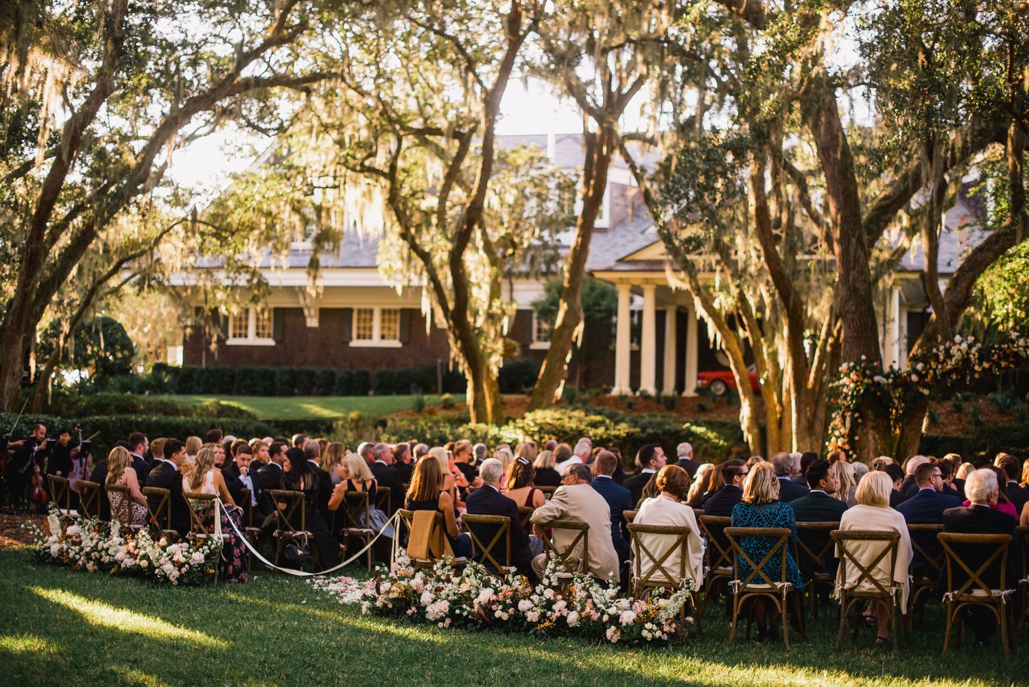 Outdoor wedding ceremony setup with chairs on a lawn surrounded by Spanish moss-draped oak trees at sunset.