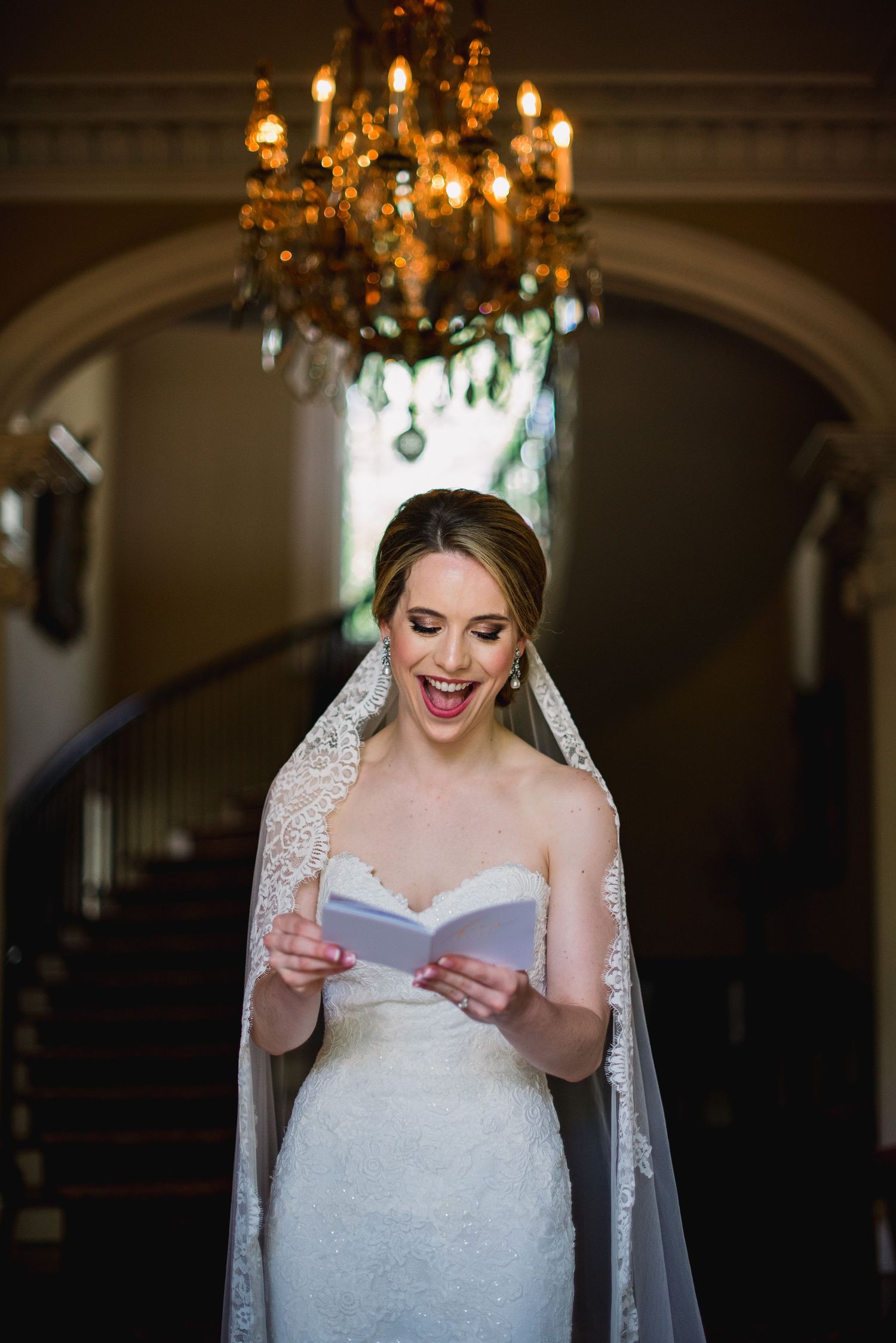 Bride in a white lace gown reads a letter with joy while standing under an elegant chandelier.