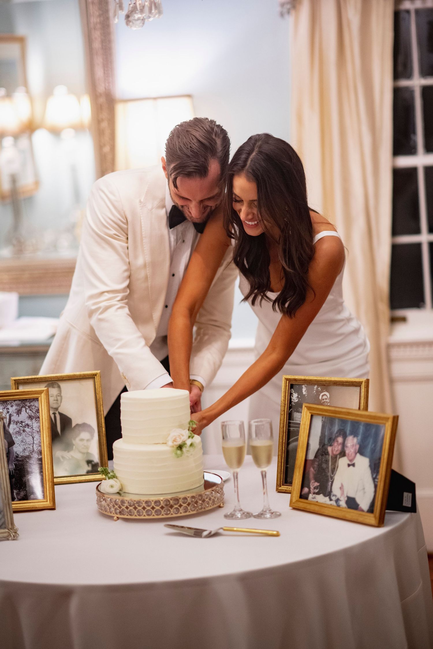 A couple in white attire cuts their elegant wedding cake together while framed photos sit on the table beside champagne glasses.