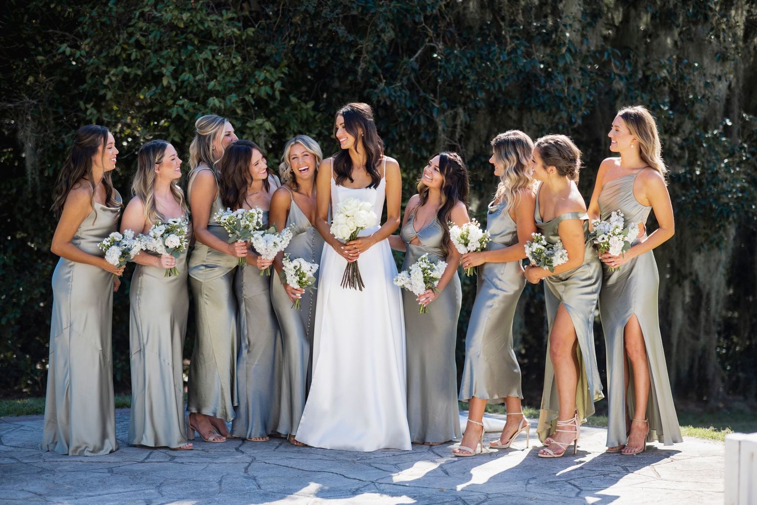 Bridesmaids in neutral gray dresses pose with bride in white gown at outdoor wedding celebration.