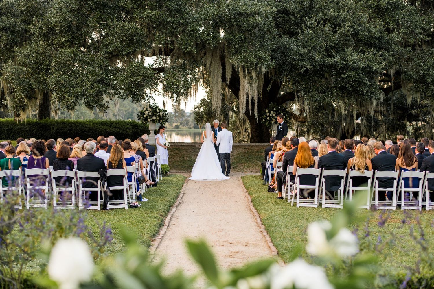 Outdoor wedding ceremony under Spanish moss-draped oak trees with white chairs and a stone pathway.