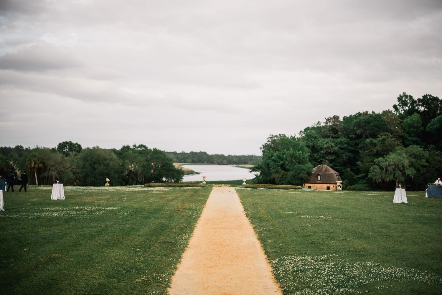 A long dirt path stretches through a green field with trees and buildings in the distance on an overcast day.