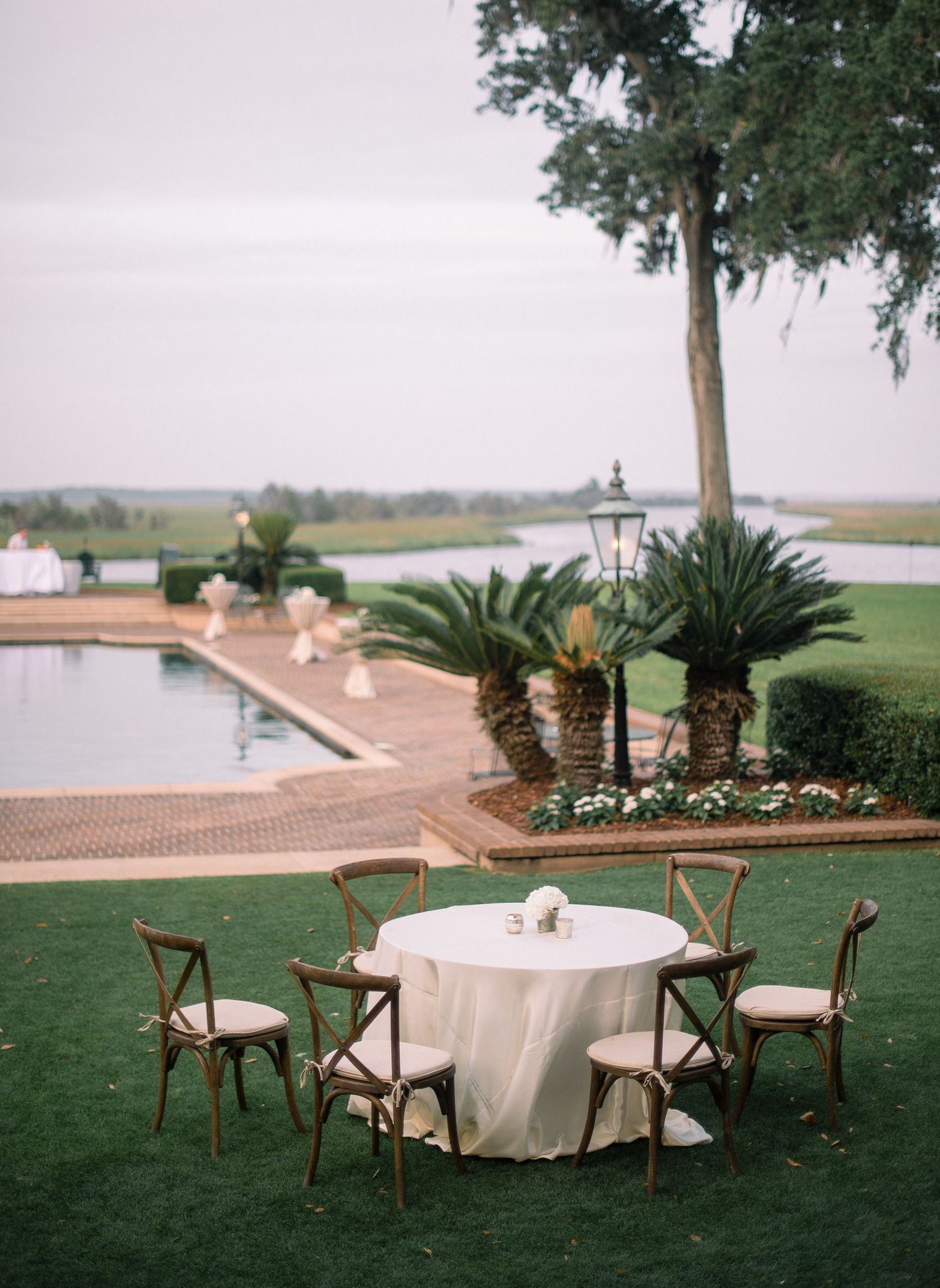 Outdoor cocktail tables with white linens and wooden chairs set up on a manicured lawn overlooking a waterfront view.