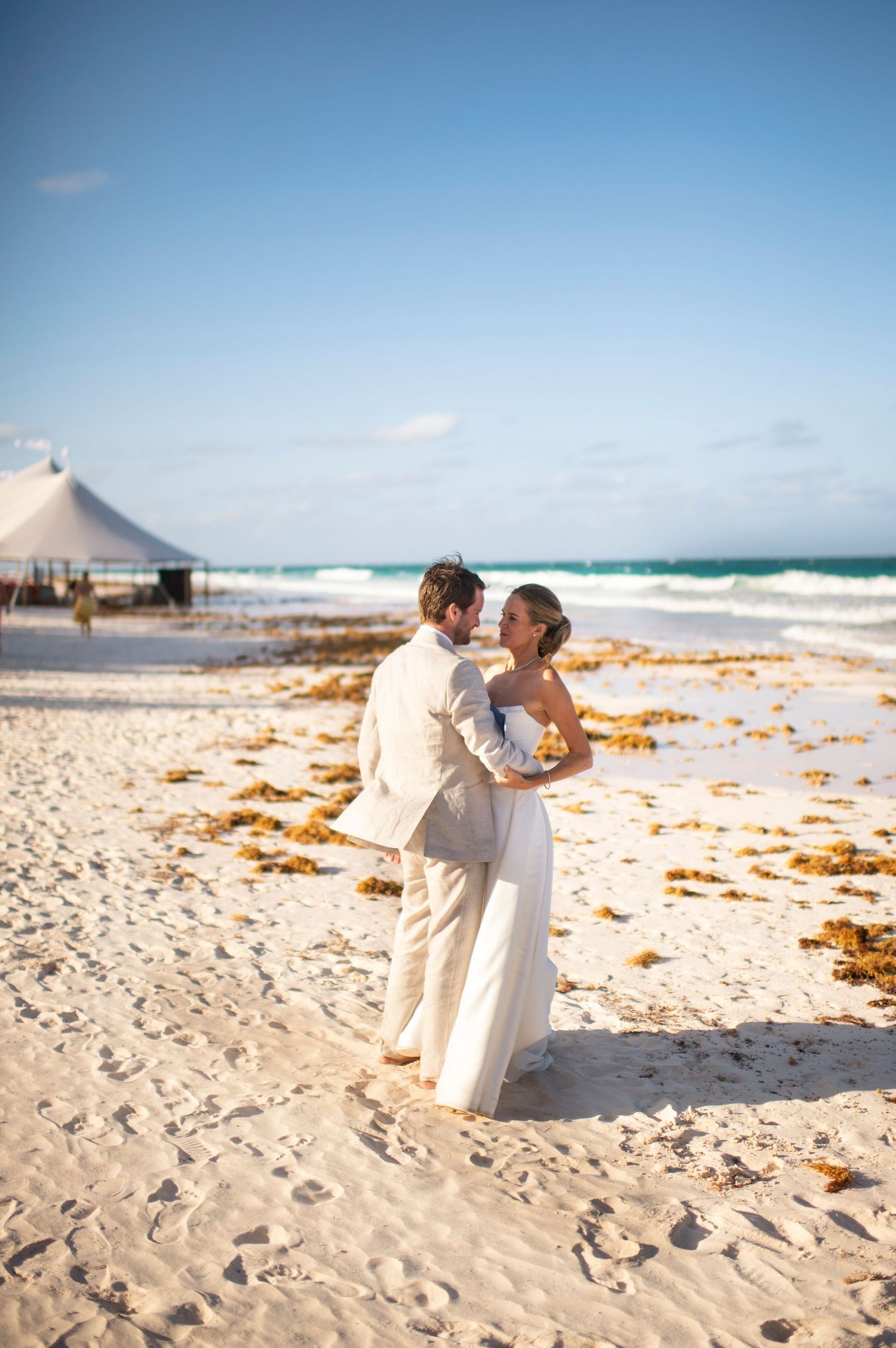 A couple in white attire shares romantic moments on harbour island Bahamas with turquoise waters and seaweed-covered sand.
