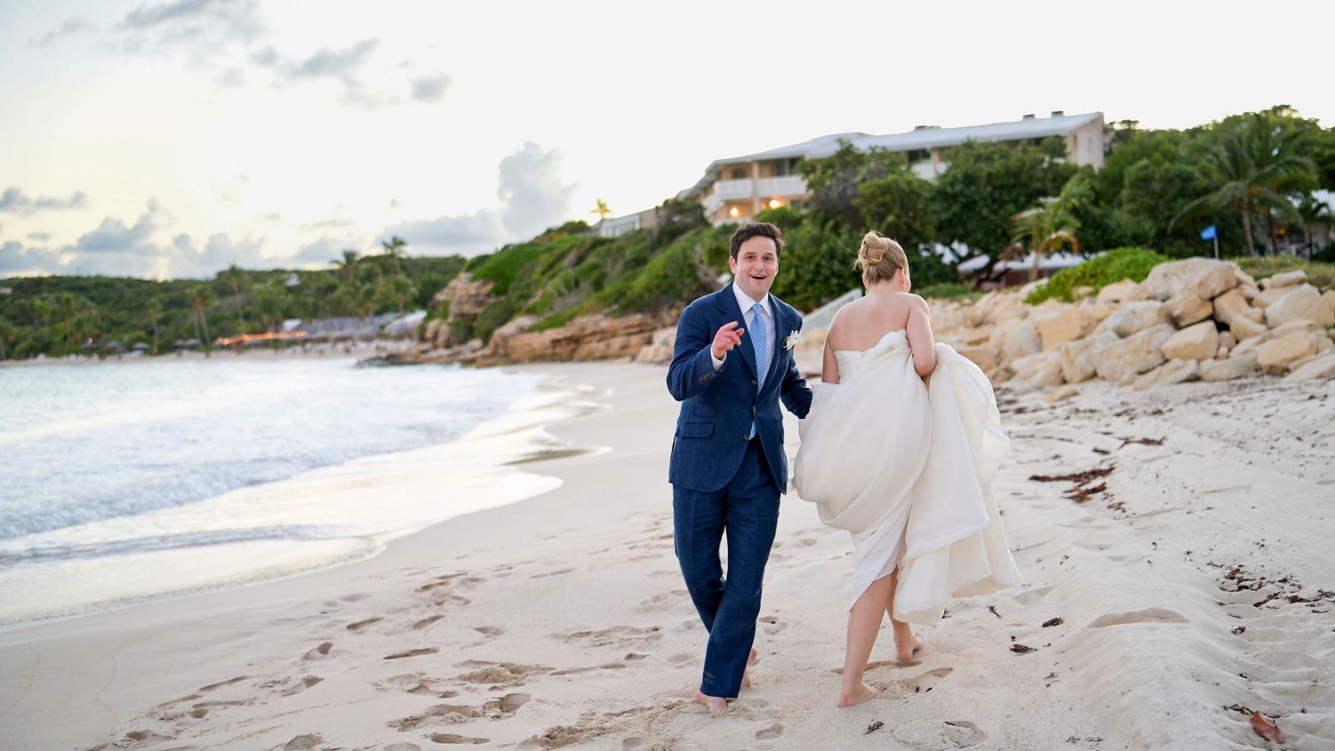 Wedding couple walking along a pristine white sand beach with coastal cliffs in background.