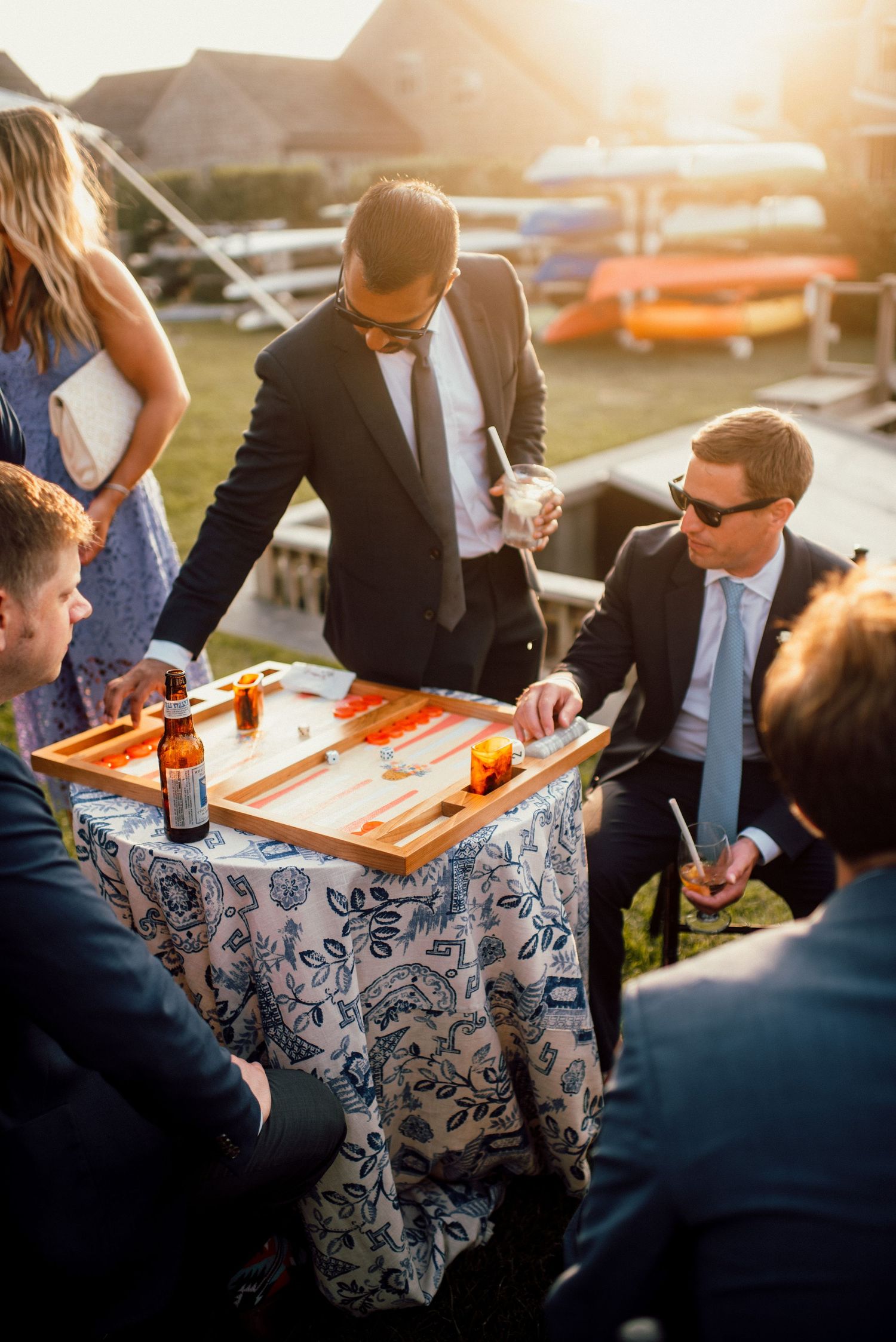 Wedding reception guests gather around tables with blue and white patterned tablecloths at sunset.