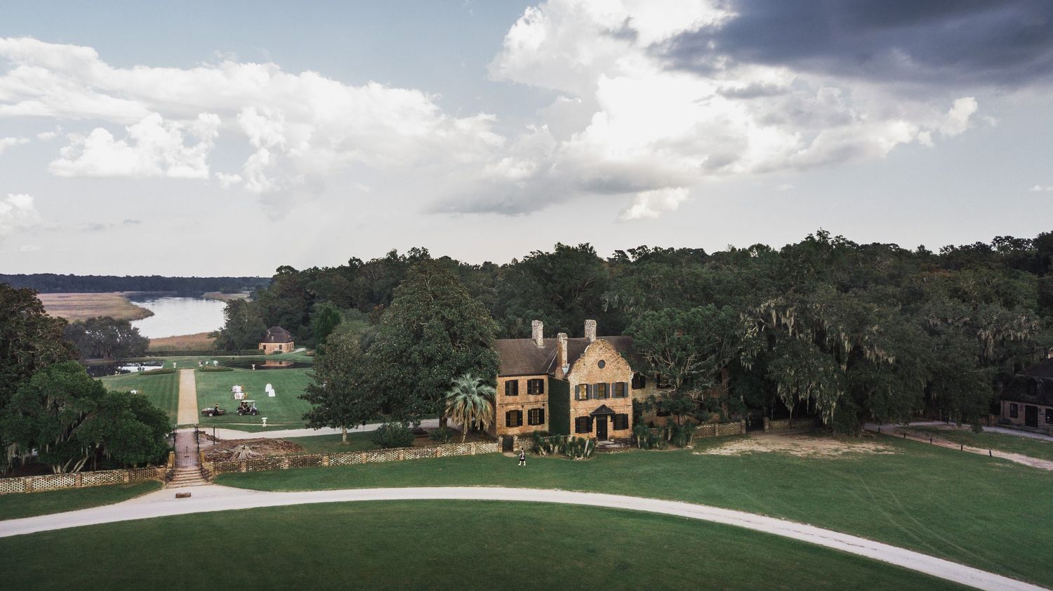 A panoramic view of a golf course with rolling green fairways under dramatic cloudy skies and trees in the distance.