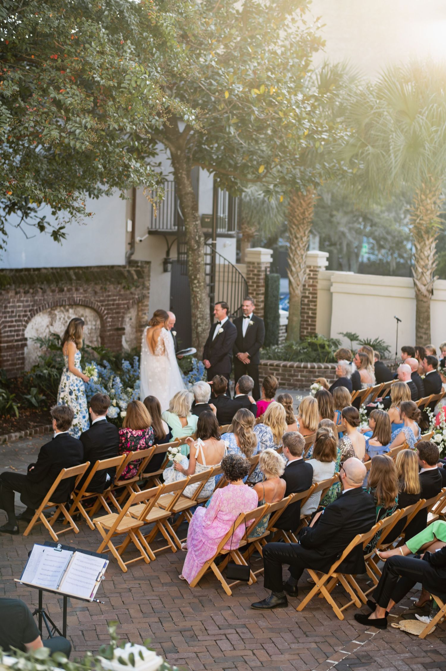 Guests seated in wooden folding chairs at an outdoor wedding ceremony on a brick patio during golden hour.
