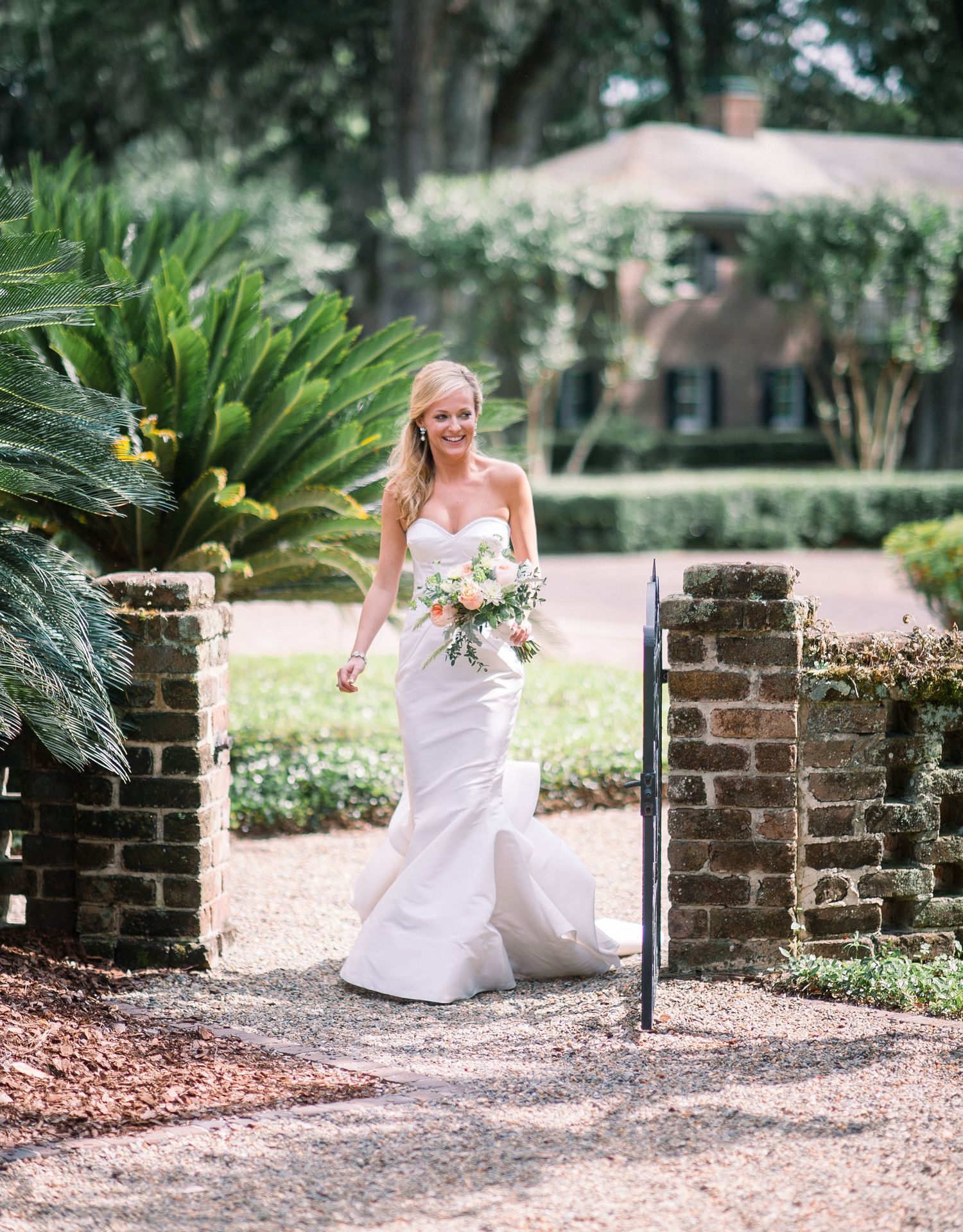 A bride in a white wedding dress poses gracefully on a brick wall path surrounded by lush greenery.