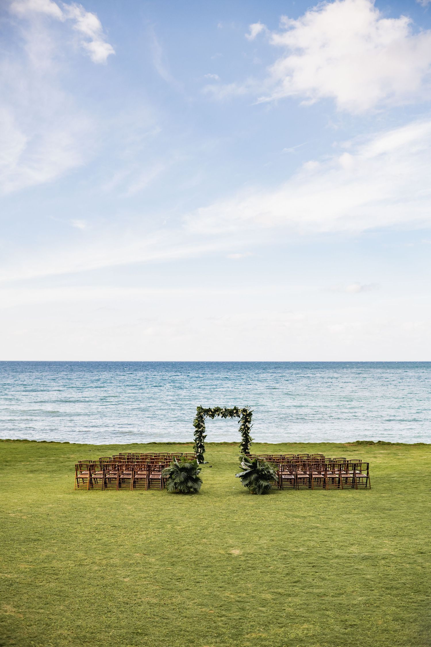 Outdoor wedding ceremony setup with wooden chairs and floral arch overlooking the ocean on a grassy lawn.