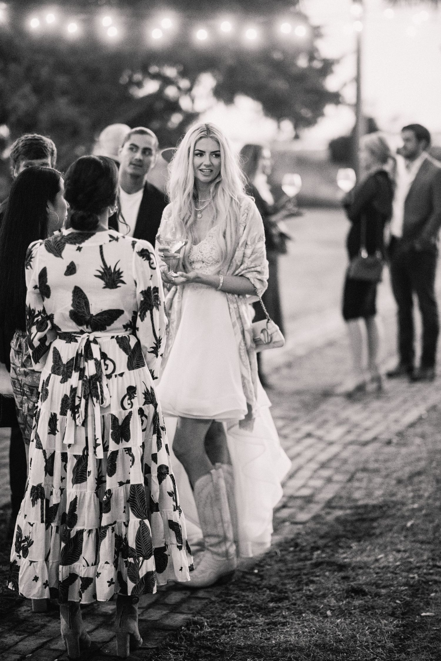 A black and white photo showing people at an outdoor gathering, with a woman in white dress talking to others.