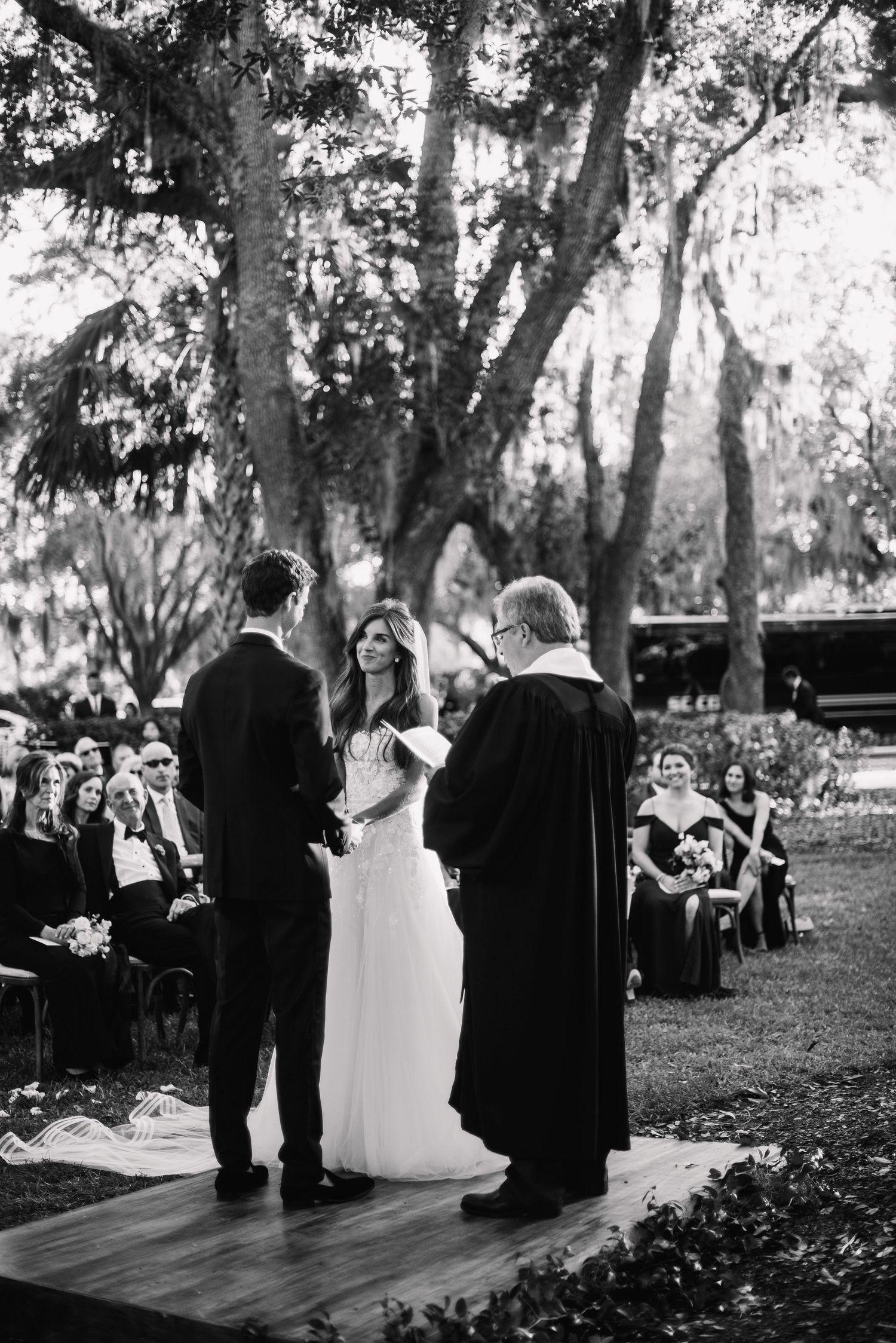 A black and white outdoor wedding ceremony takes place under tall trees with guests seated on the lawn.