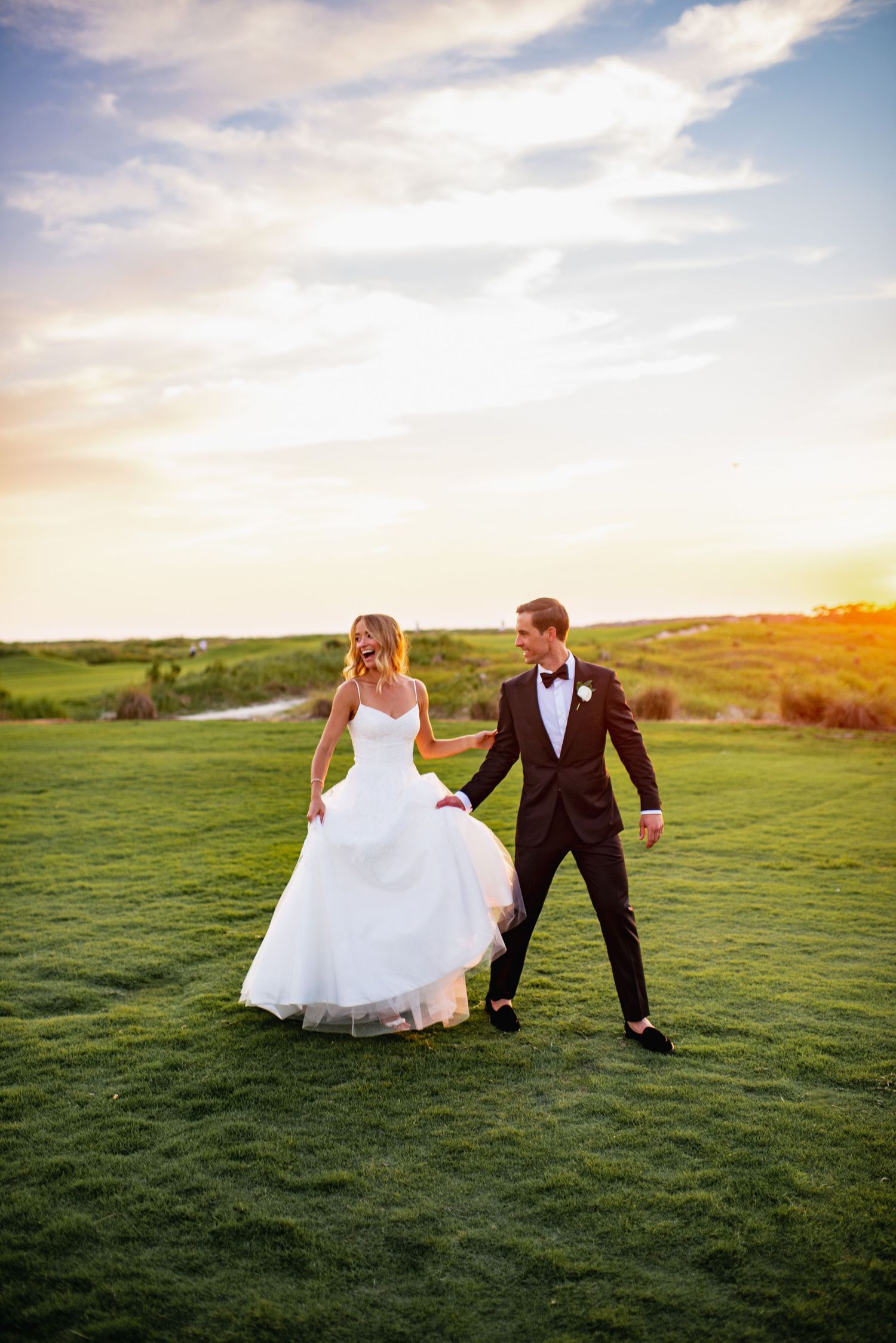 A couple in wedding attire walks together on a lush green hilltop during golden hour sunset.