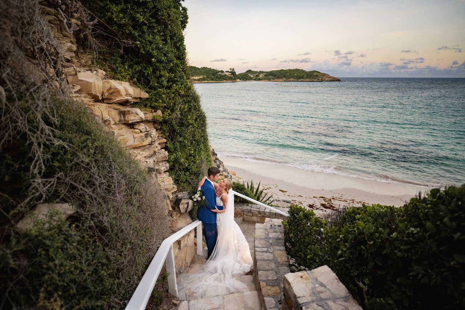 Couple in wedding attire walks along scenic coastal path overlooking beach at sunset.