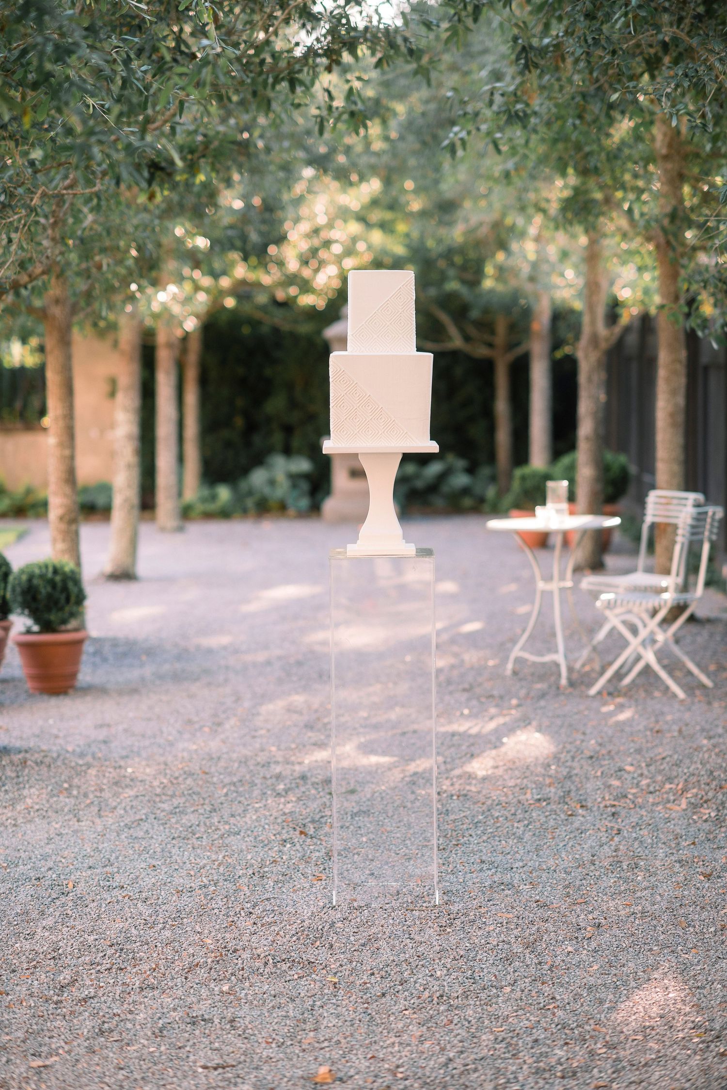 A serene outdoor patio area with white chairs and tables scattered among trees on gravel ground in soft focus.