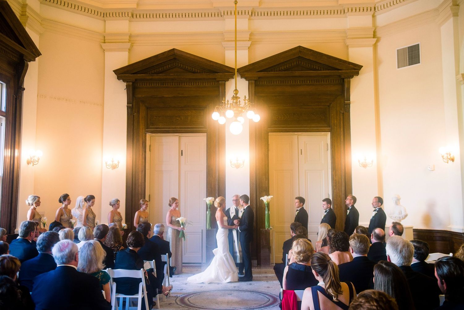 Wedding ceremony in a grand historic room with ornate architectural details and natural light streaming through windows.