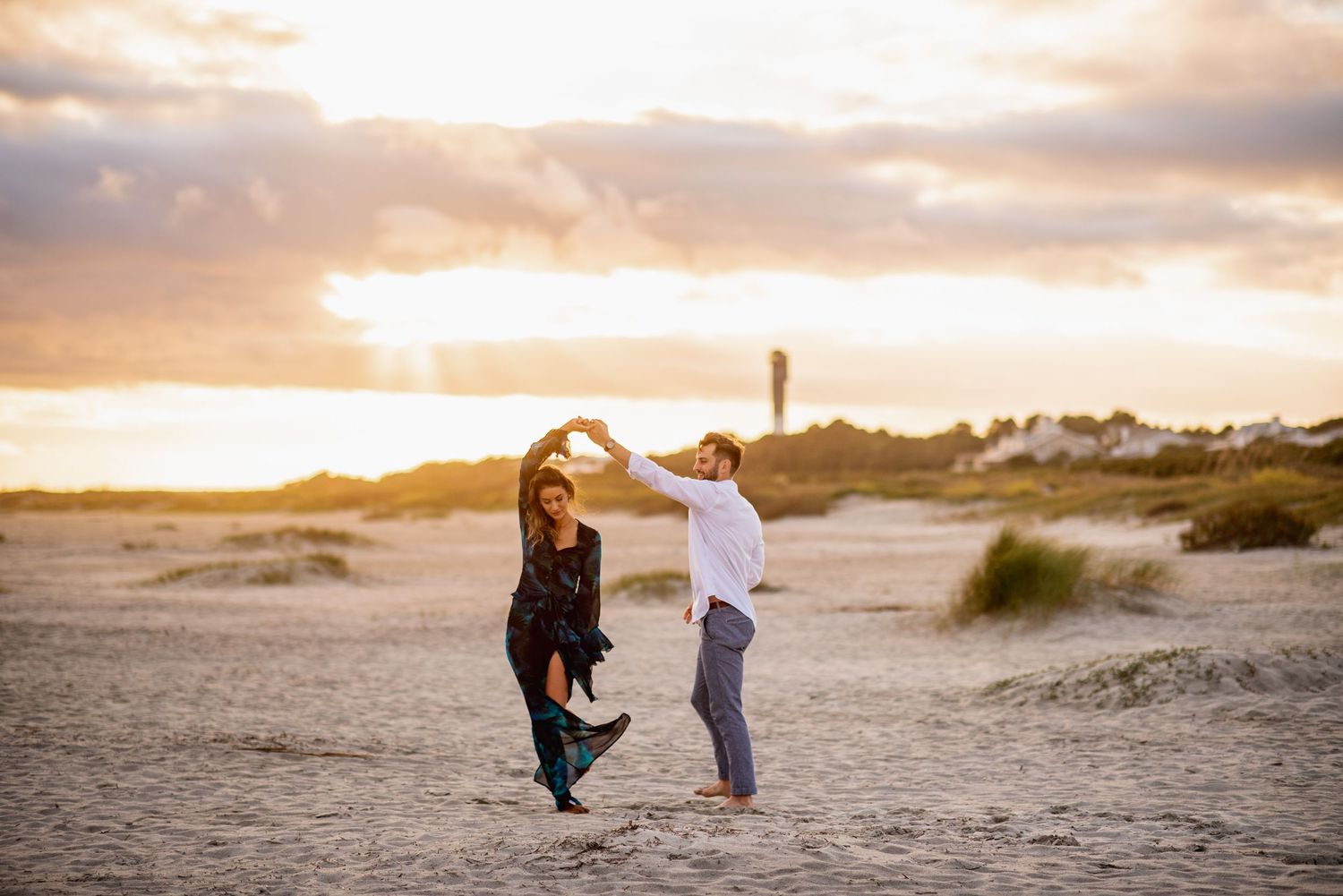 Couple walks along sandy beach at sunset with dramatic orange and pink sky in background.