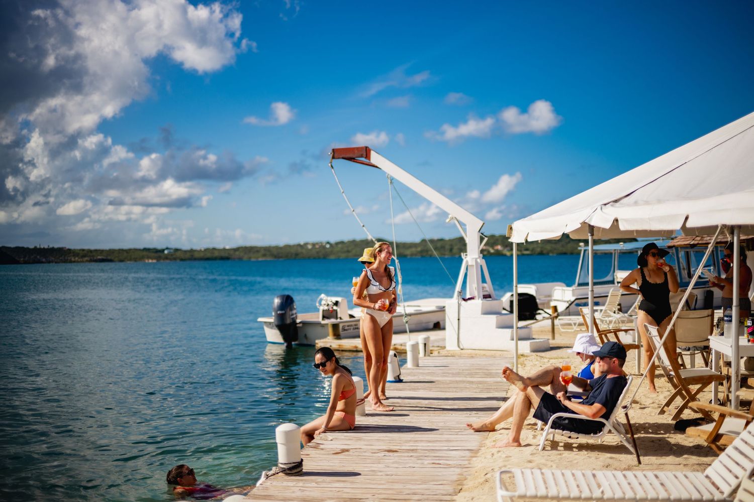 People enjoying a sunny day on a wooden dock at a tropical resort with boats and tents.
