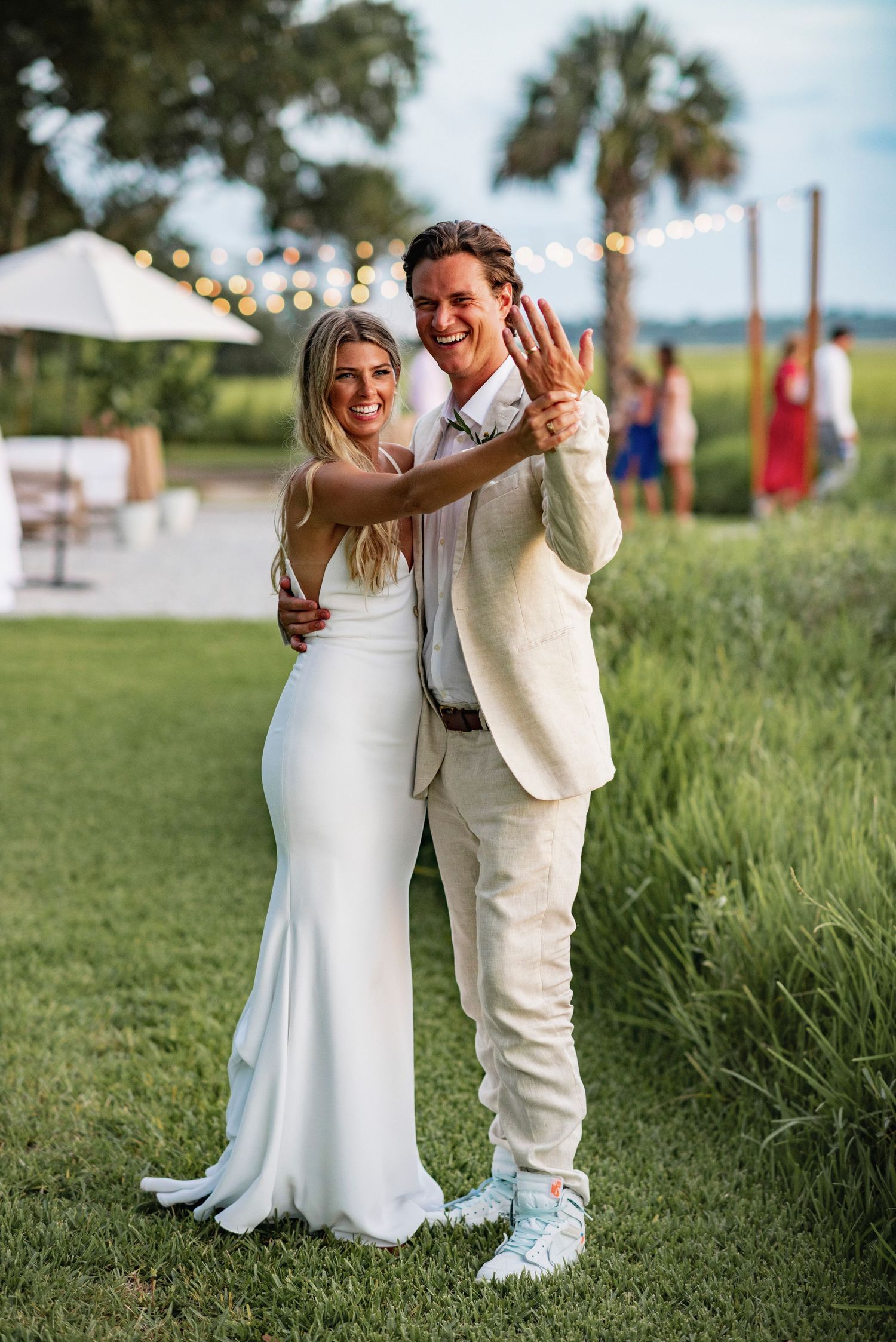 A couple in wedding attire dances together on grass, with the bride in white and partner in a light beige suit.