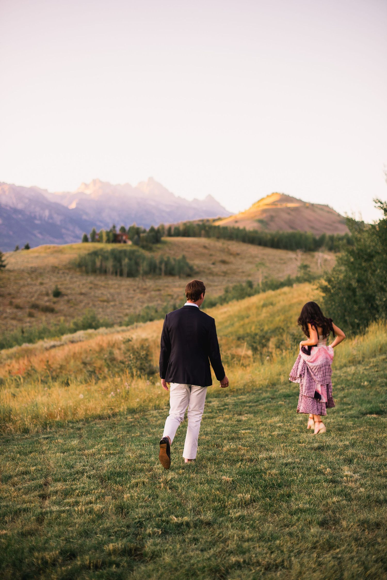 Two people walk across a grassy hillside with mountains in the background during golden hour.
