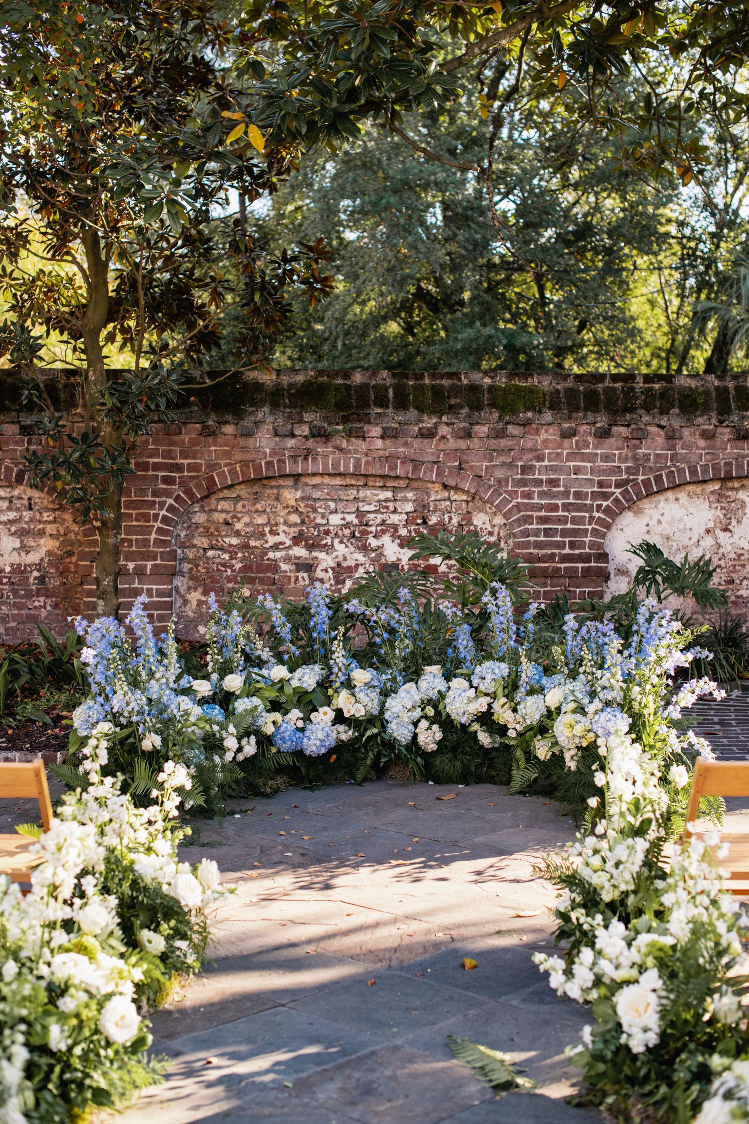 A curved brick wall adorned with elegant white and blue floral arrangements creates a romantic outdoor ceremony setting.