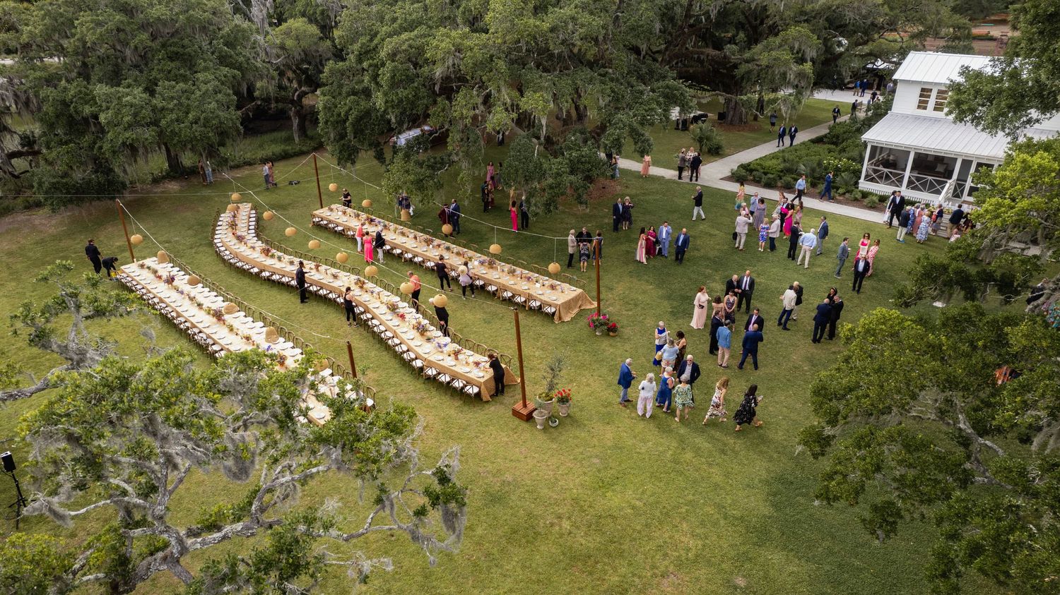 Aerial panoramic view of people gathered for an outdoor event in a lush green garden setting with winding paths.