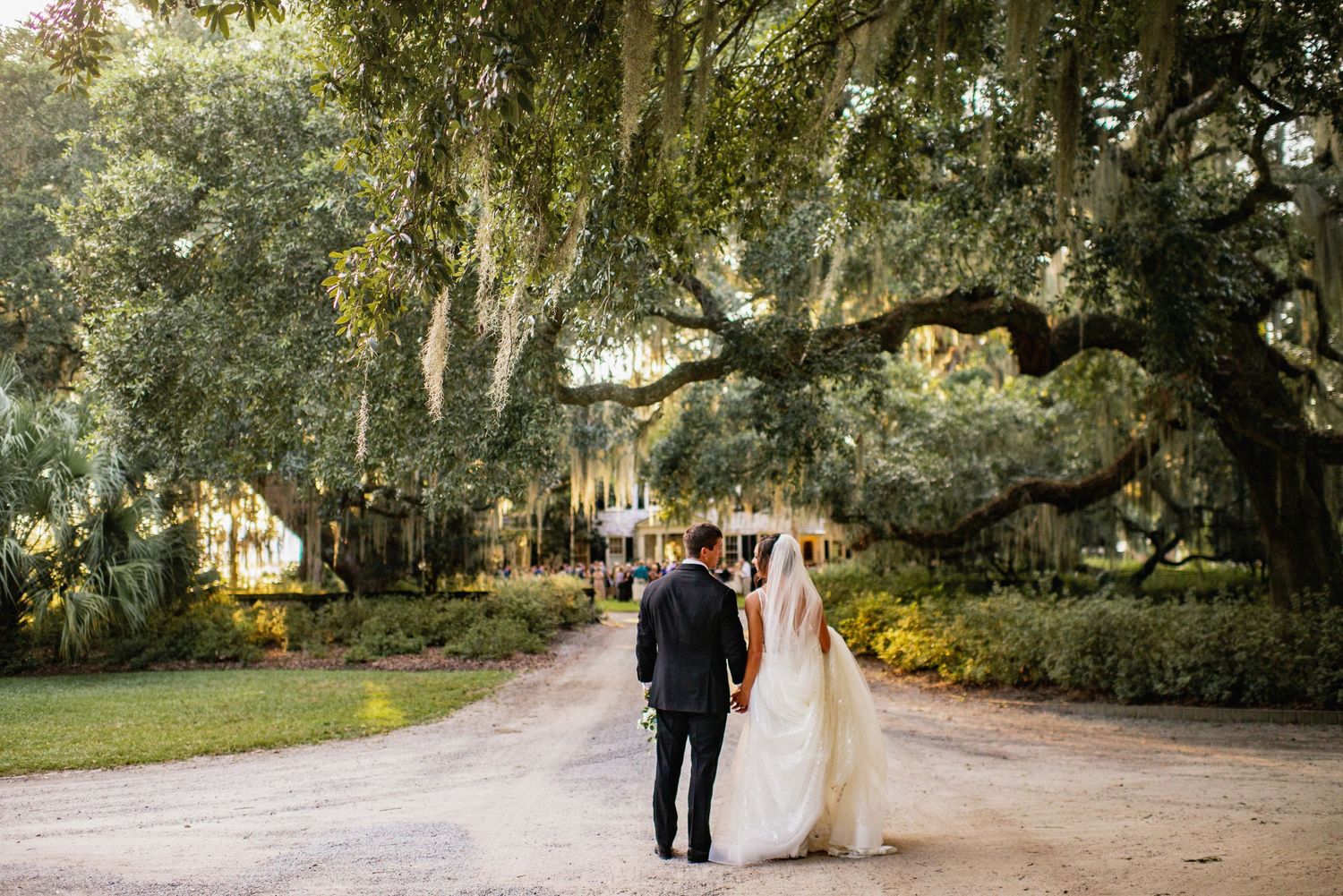 A couple walks along a romantic tree-lined path at sunset in a scenic garden setting.