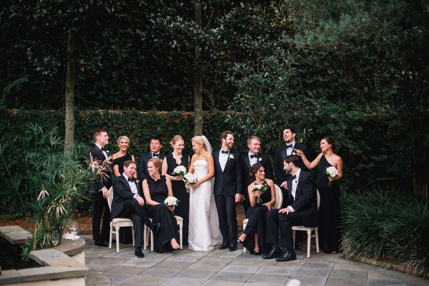 Wedding party in formal black attire poses together on stone patio surrounded by lush garden landscaping.