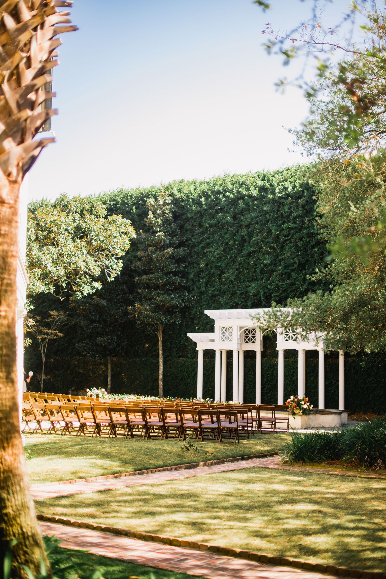 An outdoor wedding ceremony setup with wooden chairs facing a white gazebo surrounded by manicured hedges and palm trees.