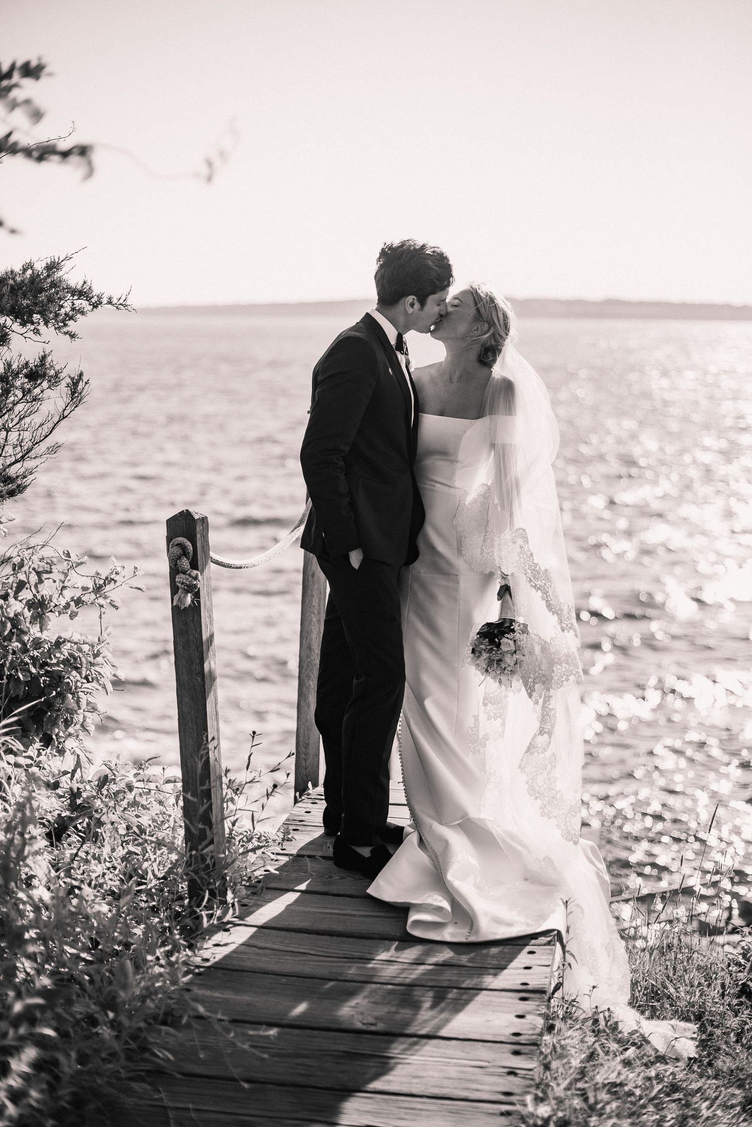 A romantic black and white wedding photo of a couple sharing a tender moment on a wooden dock by the ocean.