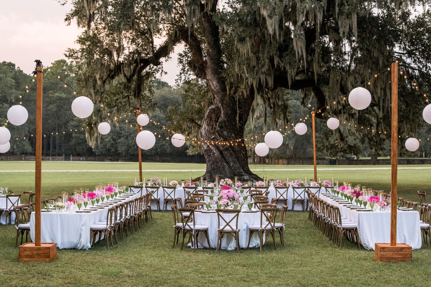 Elegant outdoor wedding reception setup with long white tables and paper lanterns under Spanish moss draped trees.