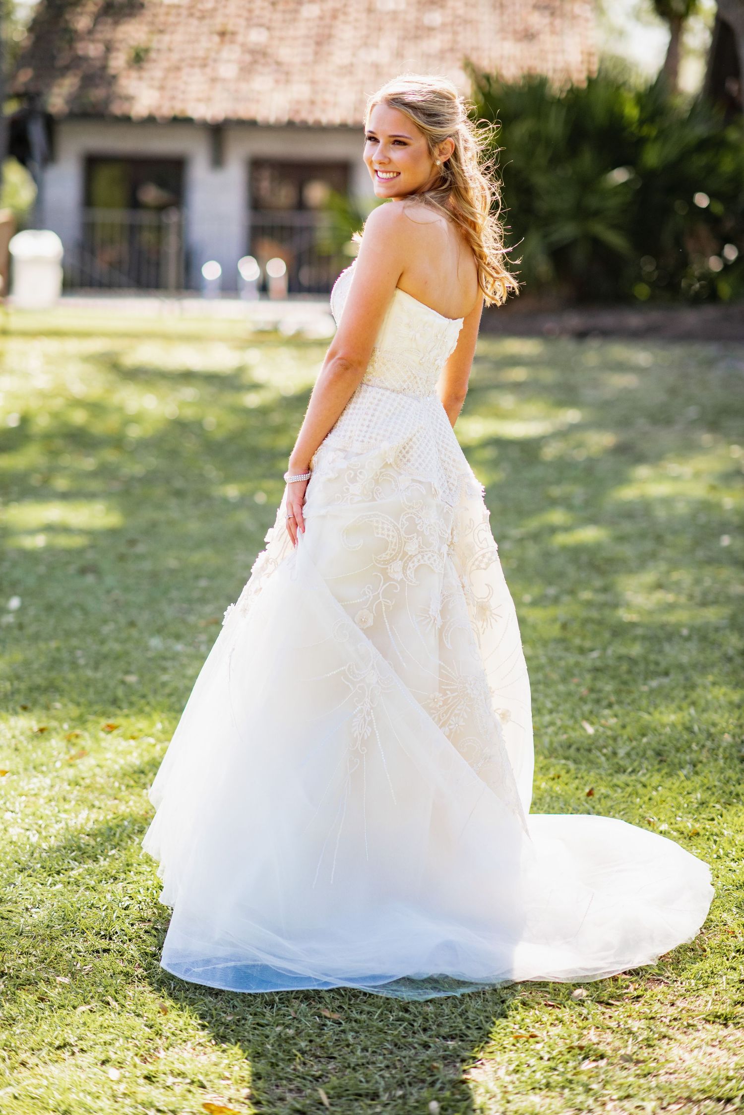 A bride in a flowing white strapless wedding gown poses outdoors on a sunny lawn with a house in the background.