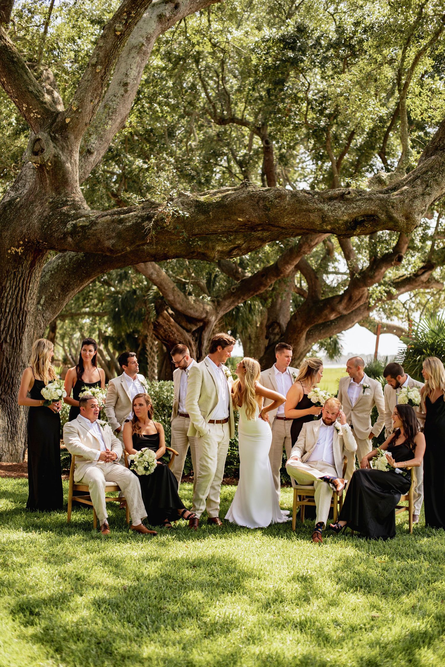 Wedding party guests in black and white attire gather under shady oak trees on a grassy lawn during an outdoor celebration.